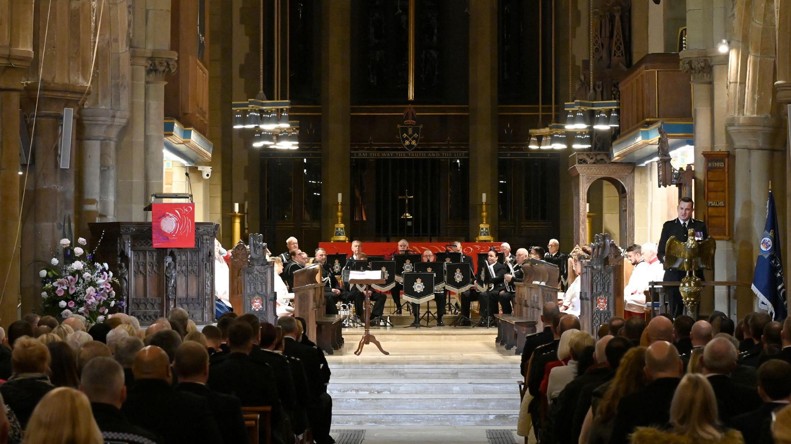 The inside of a large cathedral with high stone arches and tall columns. In the foreground, there are rows of seated people sat in pews, facing forward. They are looking on at a group of uniformed musicians seated in a semi circle, holding brass instruments.