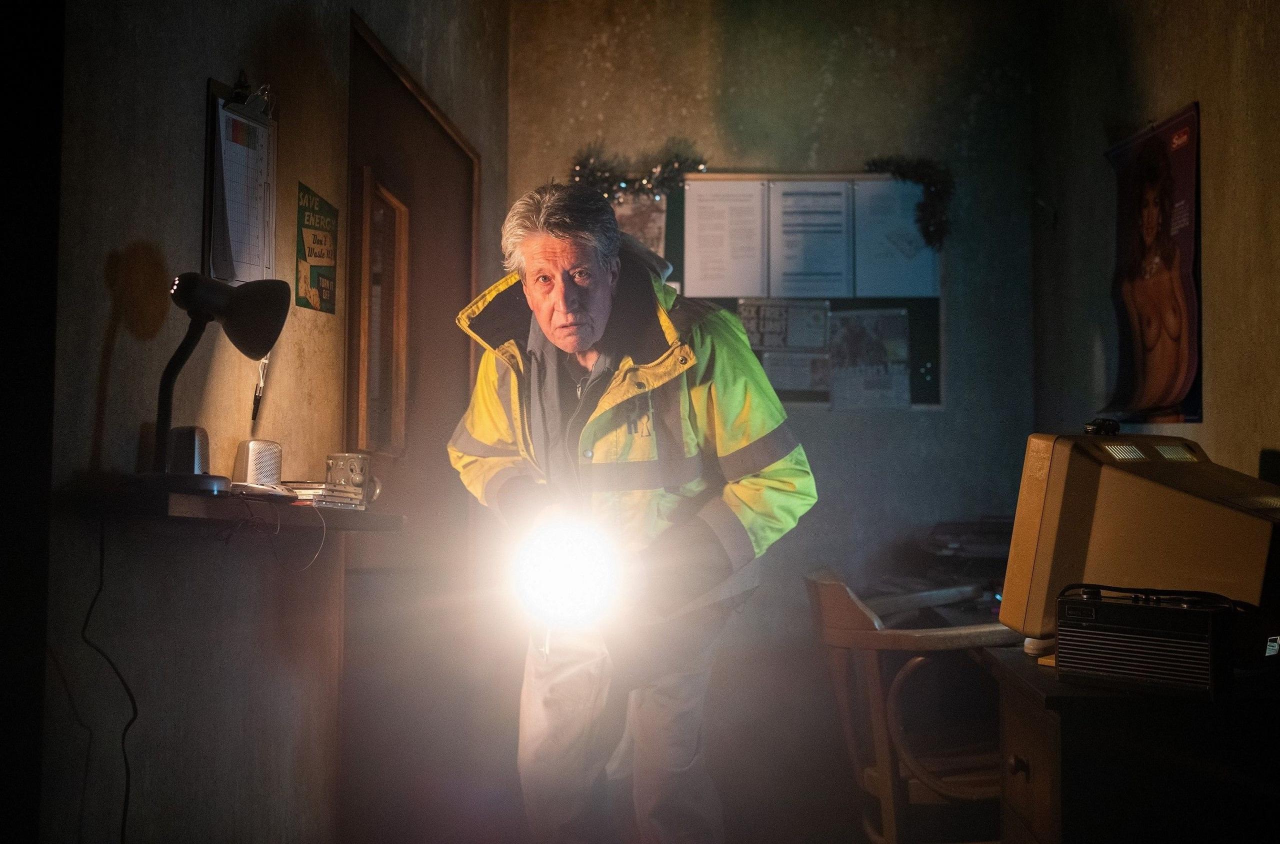 Actor David Cardy shines a torch inside a small office room during the play Ghost Stories. He is wearing a high-vis jacket and looks nervous