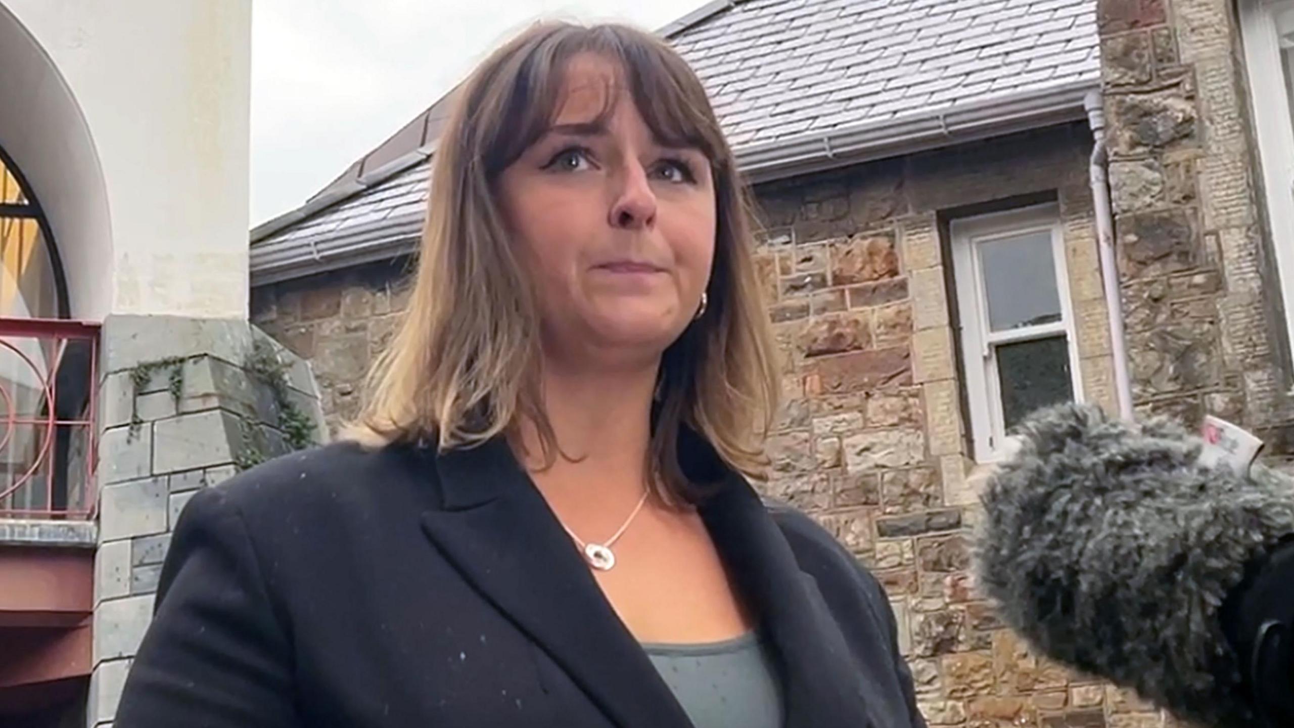 A woman wearing a dark blue suit stands in front of a building. A microphone is close to her face. 