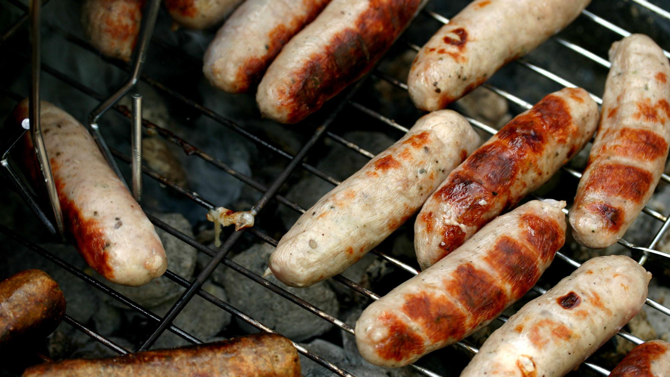 Sausages cooking on a barbecue with one being held by a tong.