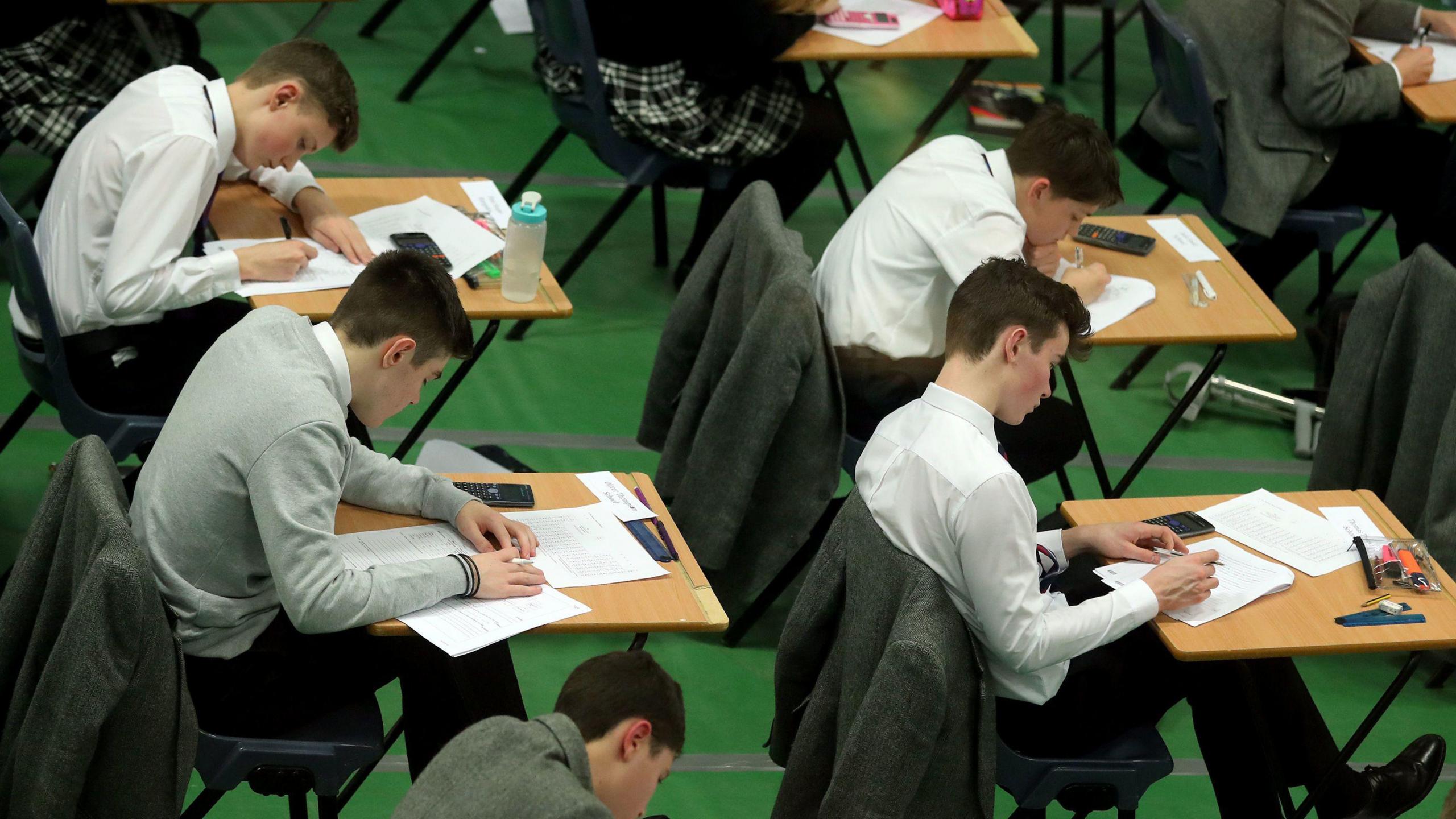 Four students take an exam while sat at small wooden tables. They are wearing white shirts with a dark school tie and light grey jumpers.