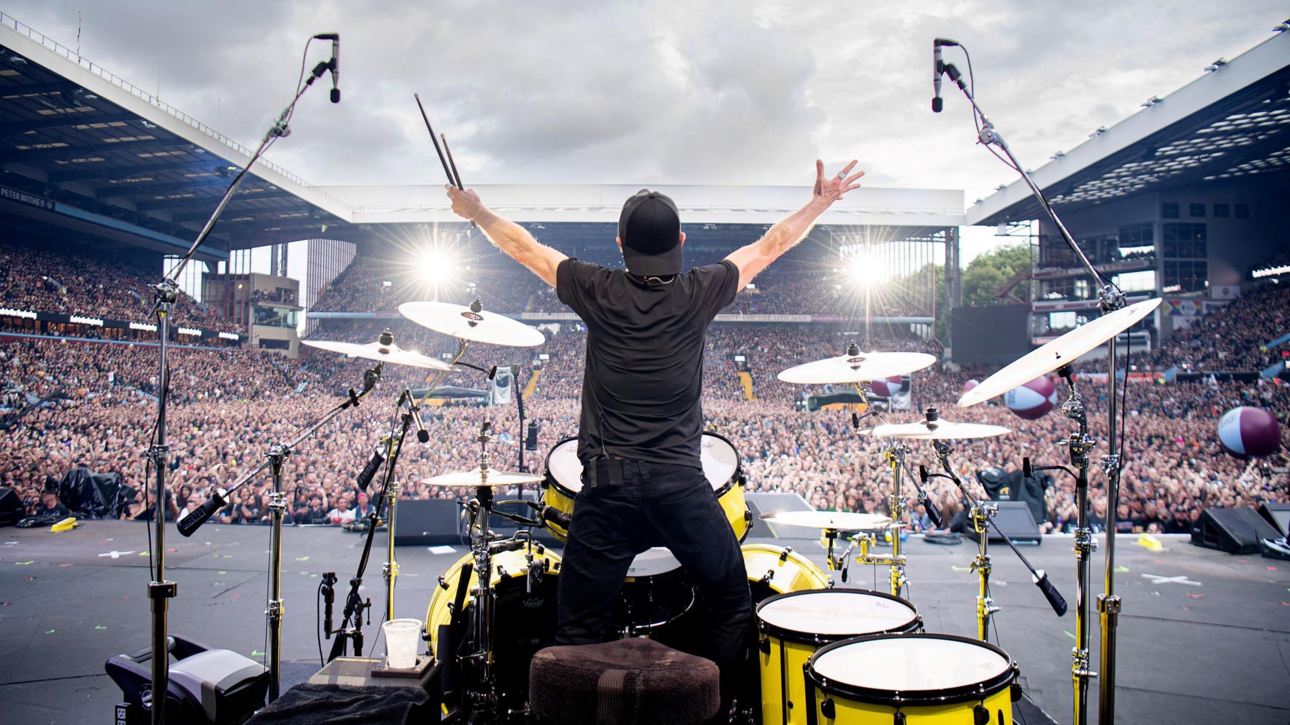 A man stands behind a drumkit on a stage and holds his arms up to a large crowd. He is wearing all black and a black baseball cap that is on backwards.