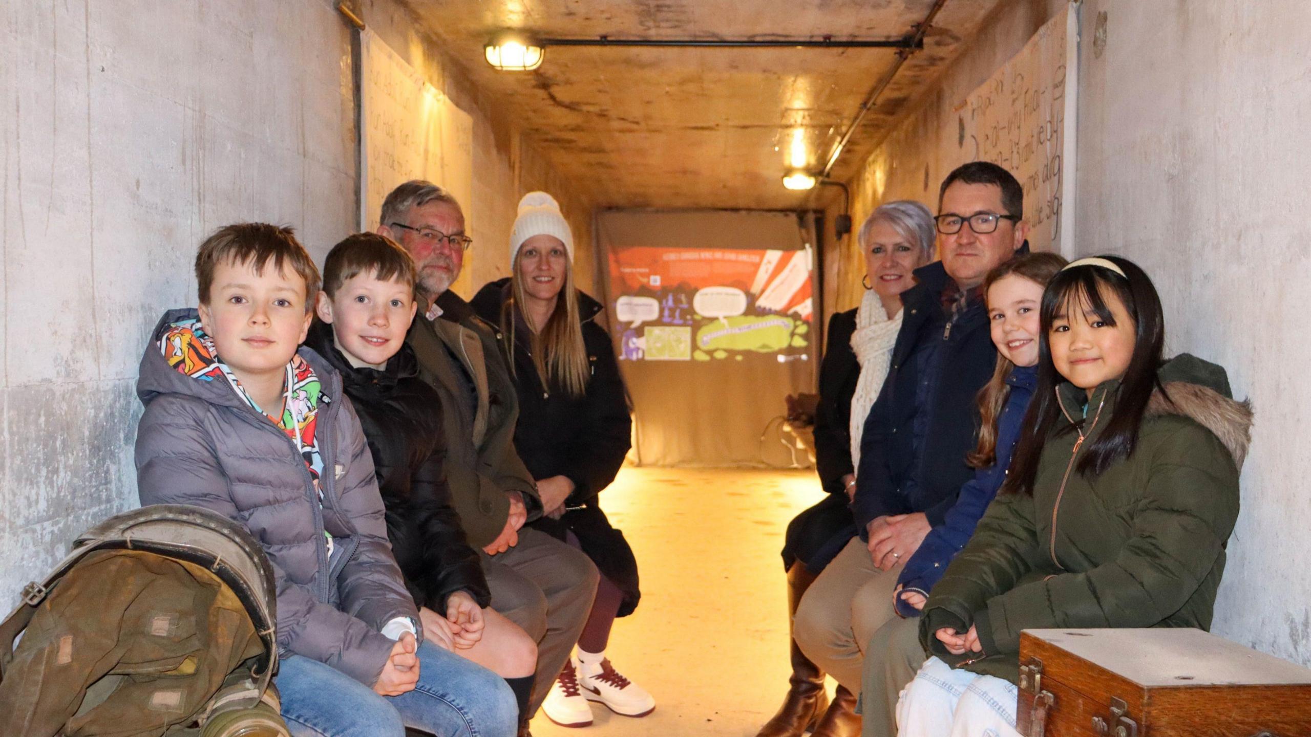 Inside a grey-coloured restored bomb shelter. Four children at the front of the image are from St Edmund Campion Catholic Voluntary Academy. Adults from left to right - Project partner Jason King, Mrs Dawson from St Edmund Campion Catholic Voluntary Academy, Councillor Abby Brennan of Rushcliffe Borough Council, Professor Chris Reynolds of NTU.