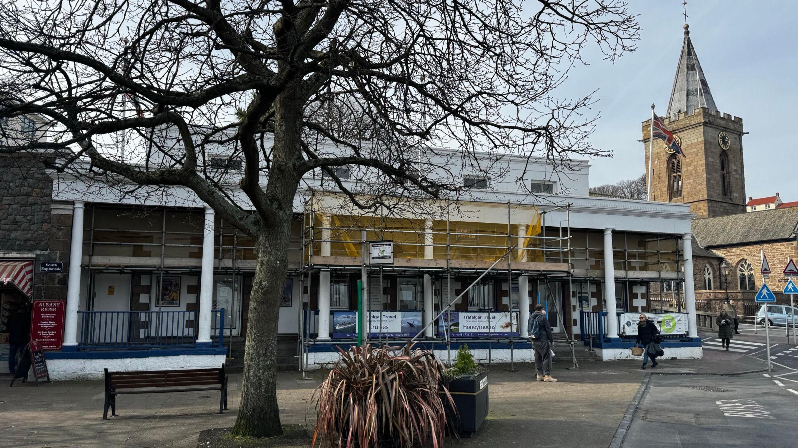 A white building with bare stone columns and window frames. One section is covered in scaffolding. Sign reads Trafalgar Travel. A tree and plant in the foreground a stone church nearby via a zebra crossing.