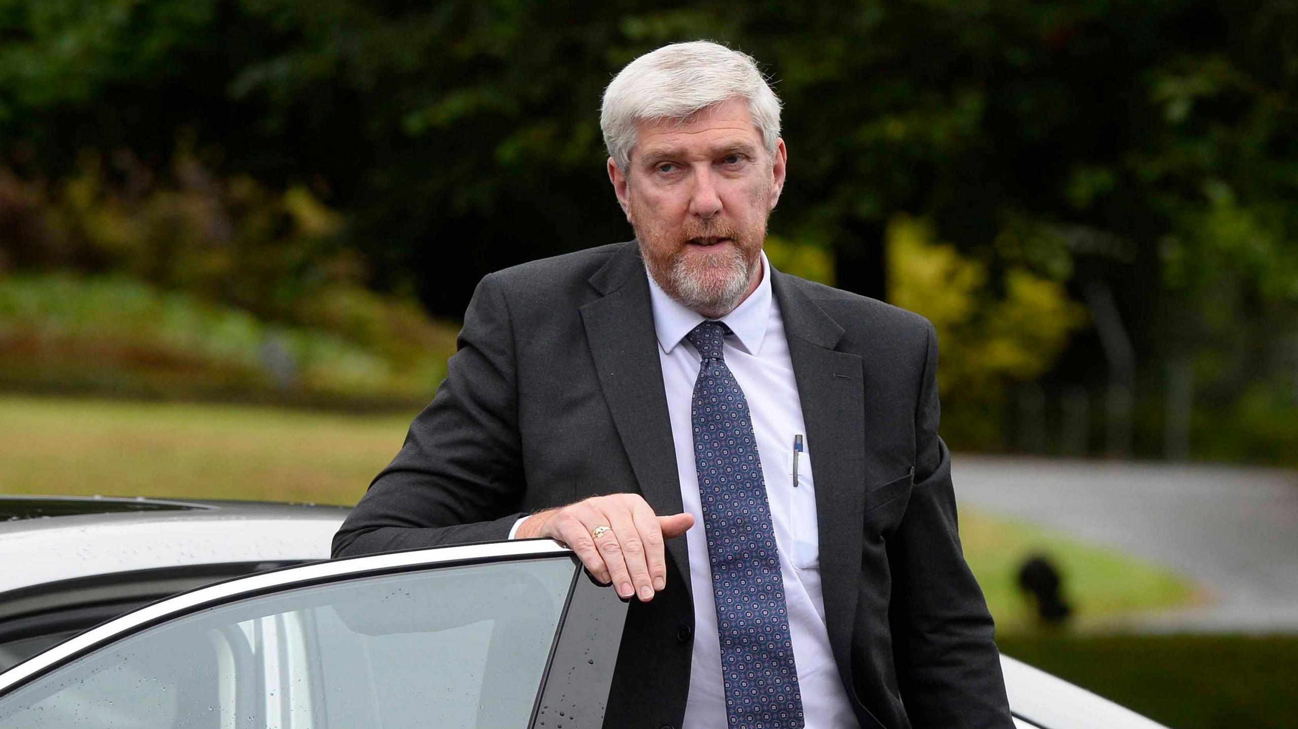 John O'Dowd gets out of a car. He has short grey hair and a grey/ginger beard and is wearing a dark suit and tie and a light blue shirt