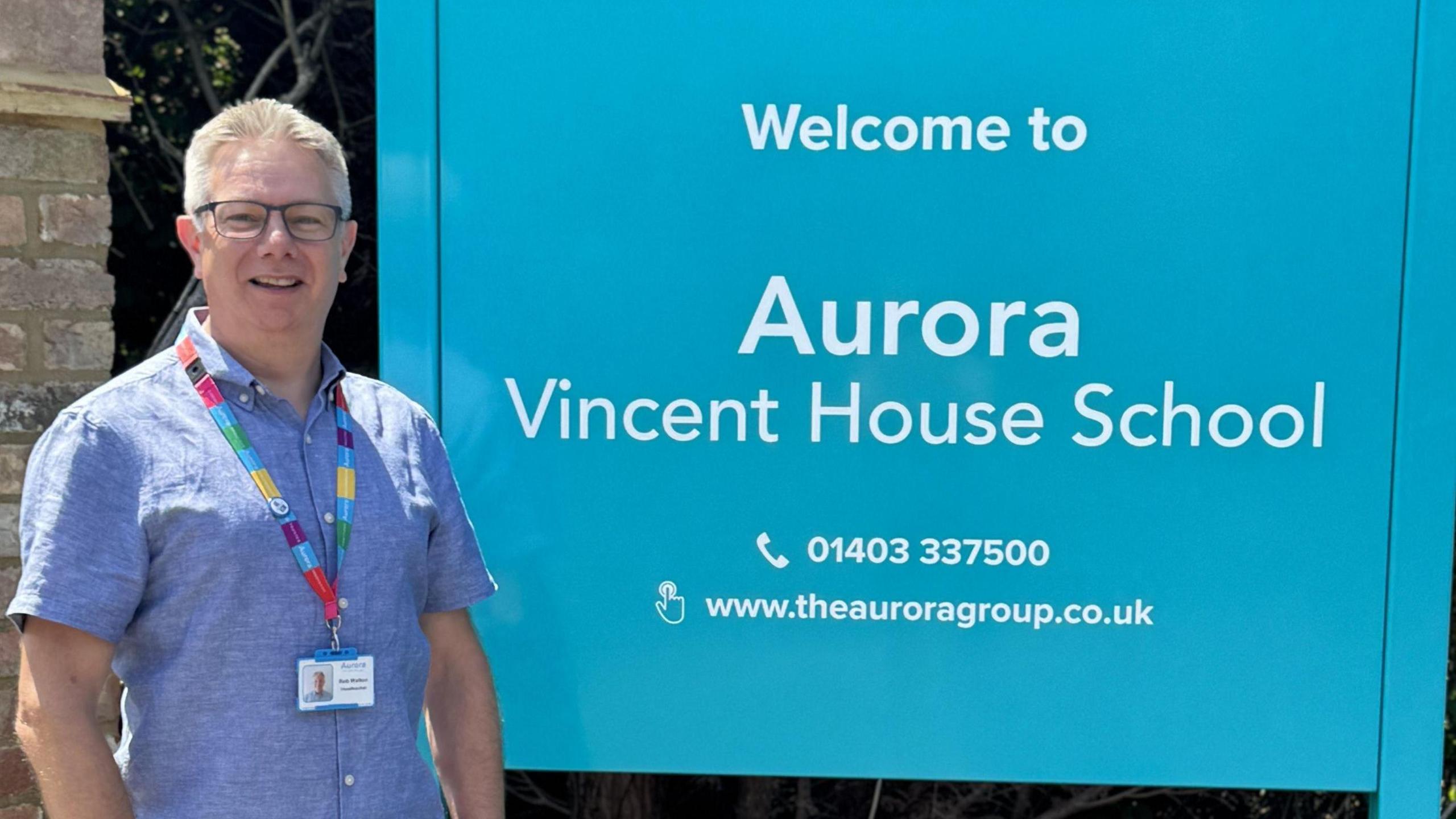 A man in a short-sleeve, blue, button-up shirt standing in front of a blue sign which says "Welcome to Aurora Vincent House School".