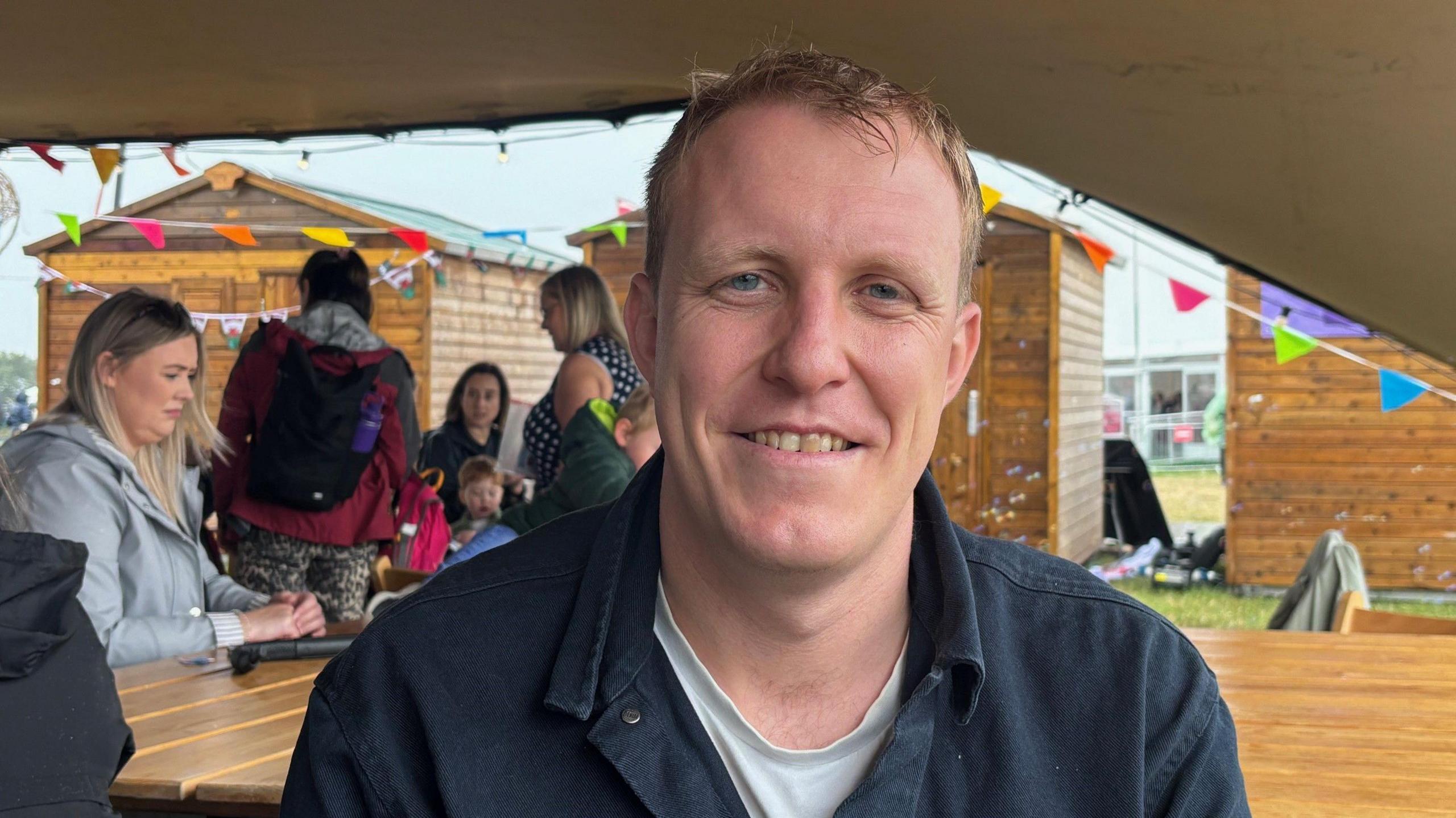 Dr Steffan Evans, smiles at the camera underneath a tent. Colourful bunting can be seen behind him. He has blue eyes and strawberry blonde short hair. 