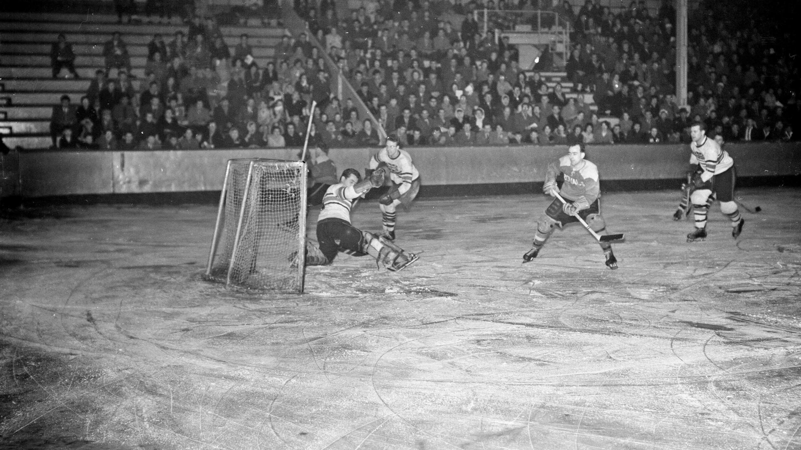 A hockey game at Durham Ice Rink in 1961. The audience is looking on as players are skating. They chasing the puck and attempting to score.