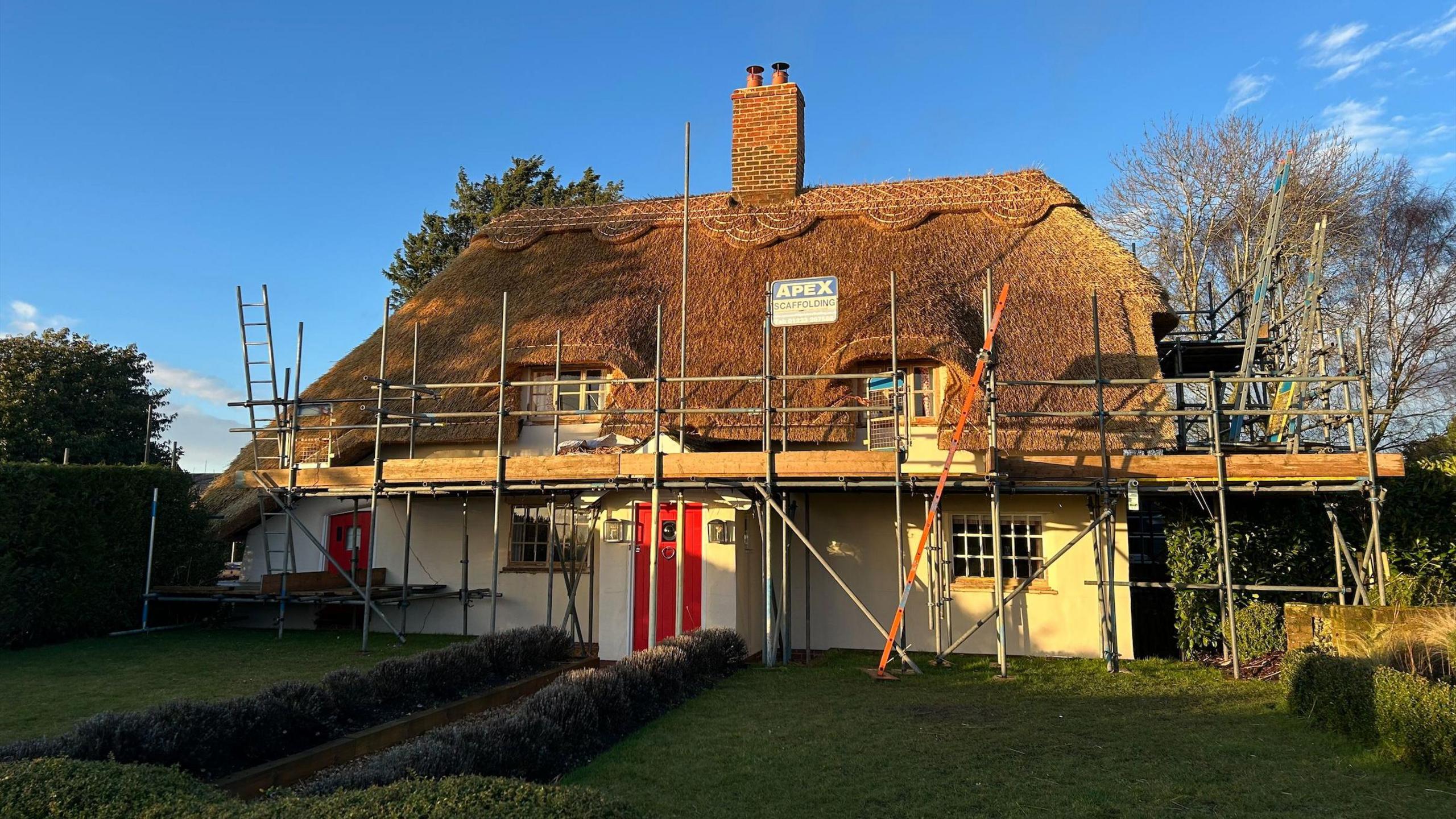 A white cottage with a red door that has a thatched roof. The house has scaffolding all around it. There is a blue sky behind the building and a garden in front. 