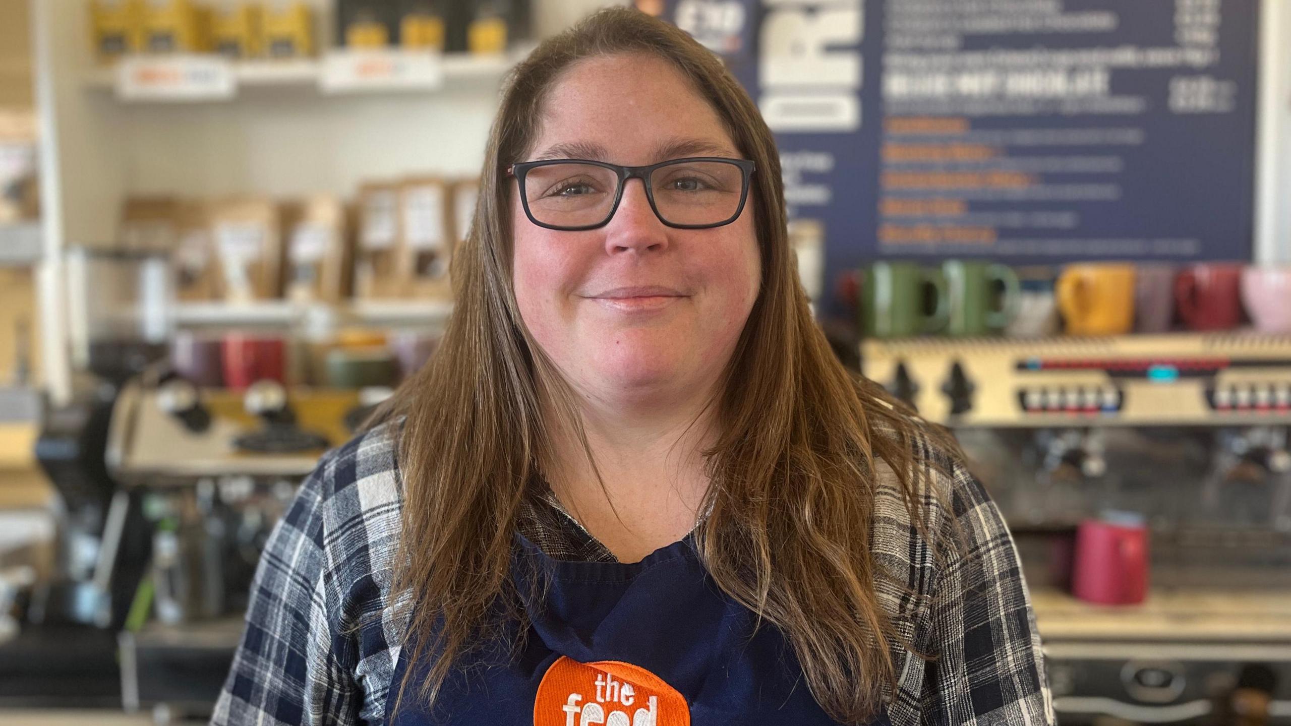 Grace Stewart has long brunette hair. She is wearing a blue apron which says "The Feed" in white writing on an orange circle. She is wearing a blue checked shirt and black-rimmed glasses. She is standing behind the counter at a cafe. She is looking at the camera and smiling.