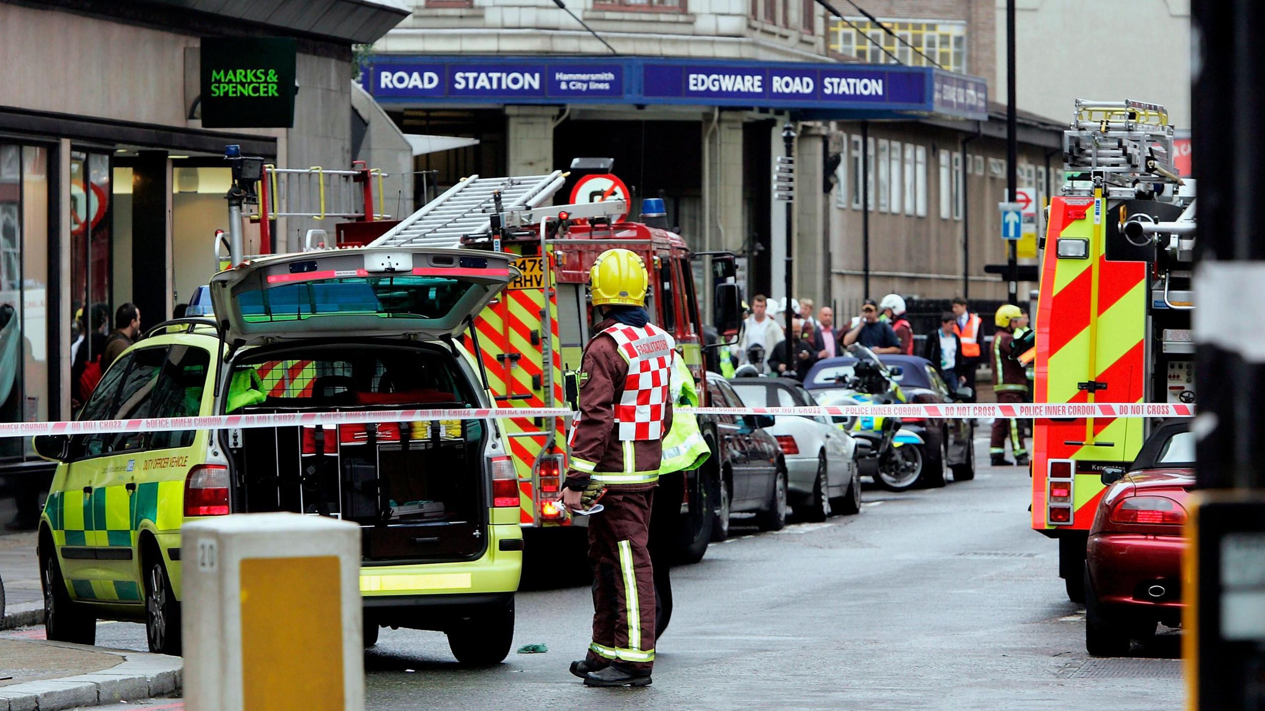 Emergency response scene outside Edgware Road Station with multiple fire engines, an ambulance, and emergency personnel. A firefighter in a yellow helmet and reflective gear stands near the vehicles. The area is cordoned off with tape, and there are several onlookers in the background.