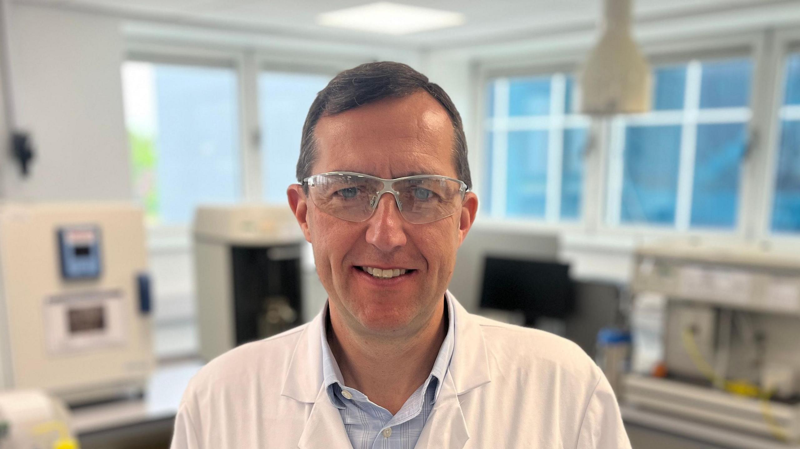 Chris Morgan in a white lab coat looking at the camera through safety glasses in a lab