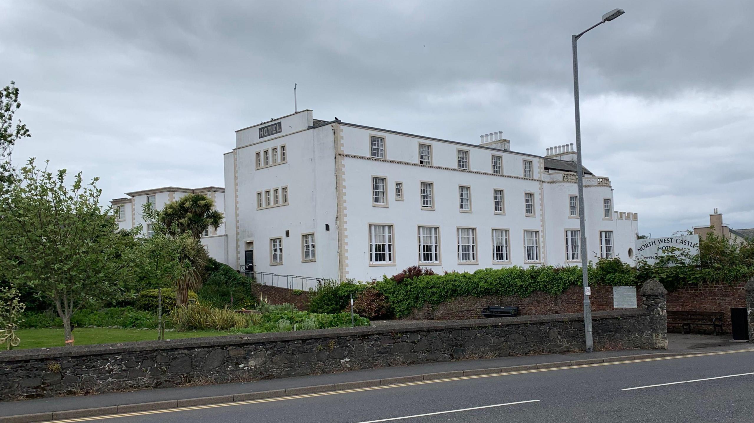 A white fronted hotel with a large number of windows behind a low stone wall and trees with the sign North West Castle Hotel almost obscured by plant life