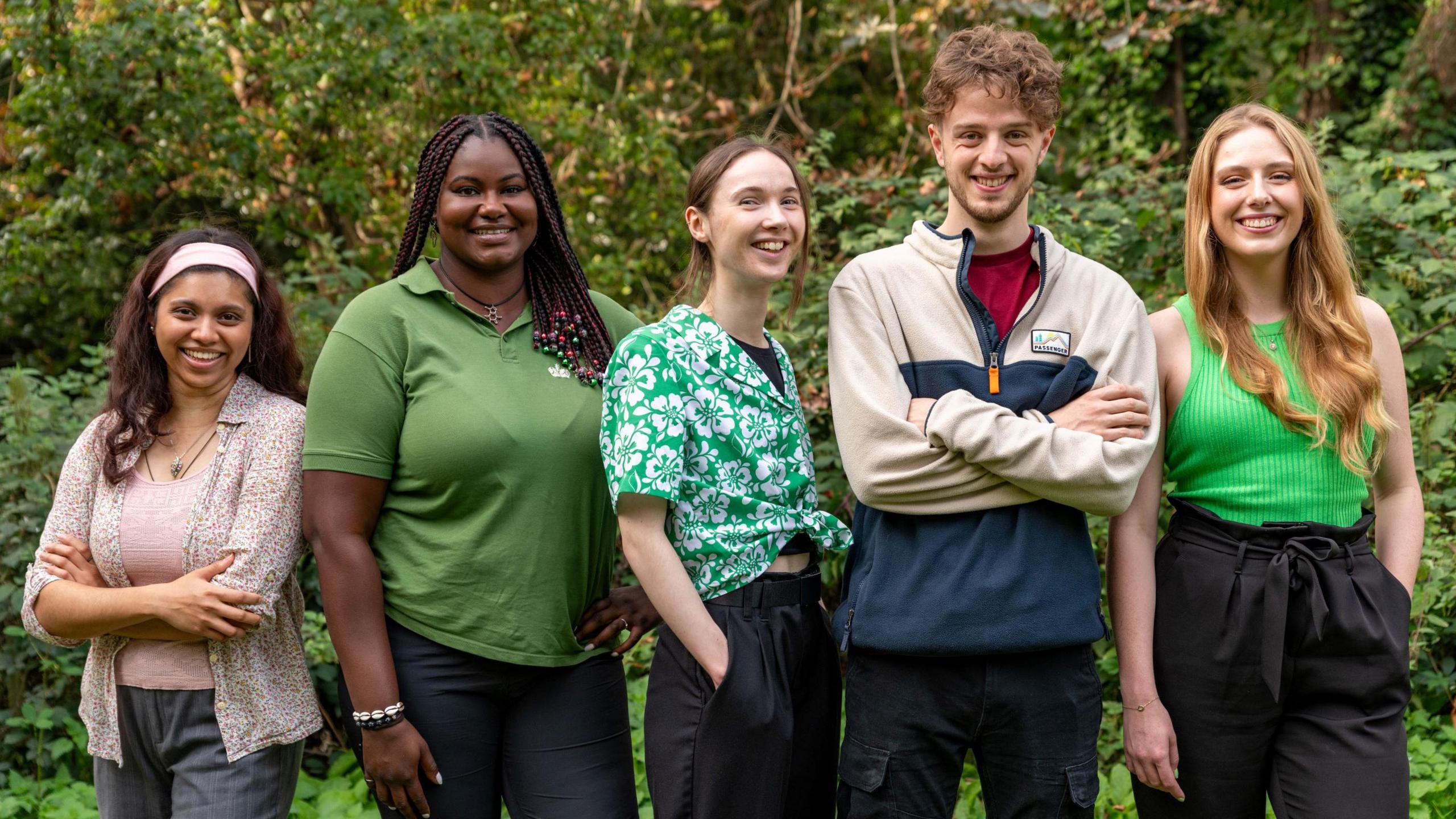 A group of five young people, one male and four female, stand in a line and smile as they pose for the camera. Three of them are wearing green tops and behind them is a bright green hedge.