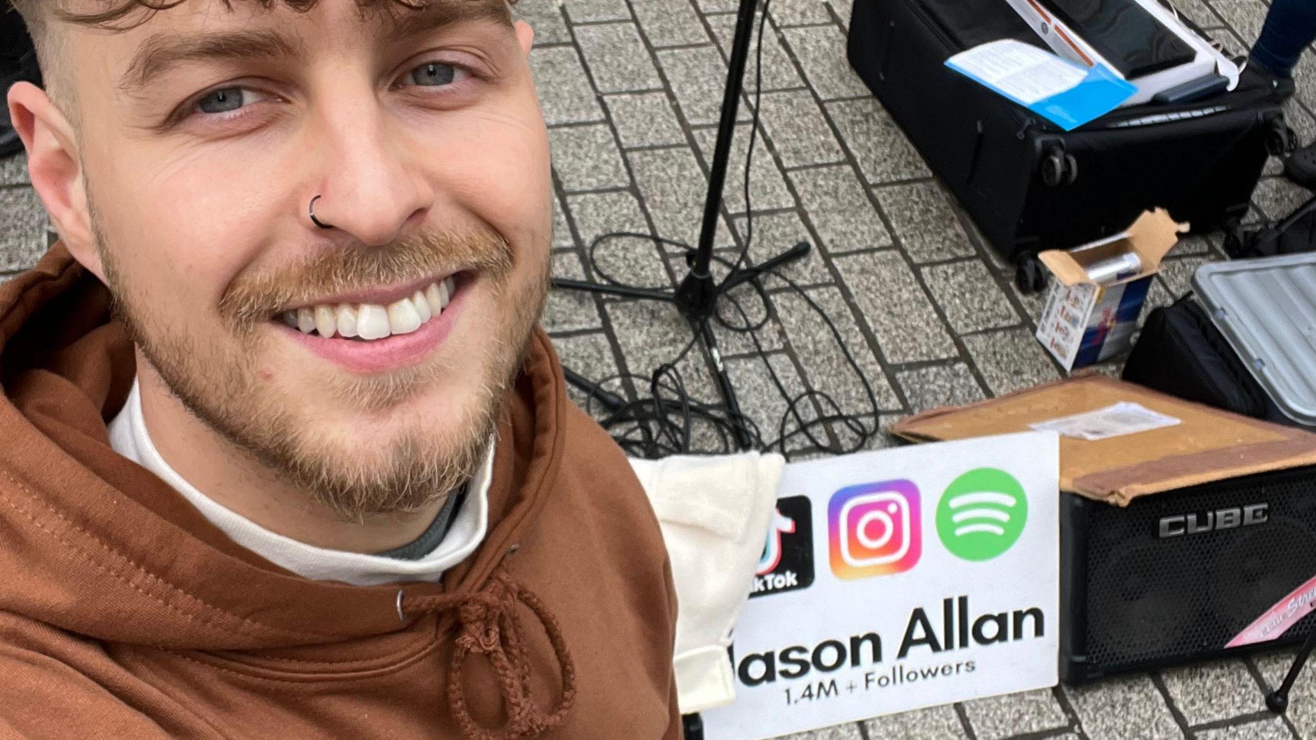 A man with brown hair and a small beard, with a black nose ring, wearing a brown hoodie smiles at the camera. He is standing next to music equipment.