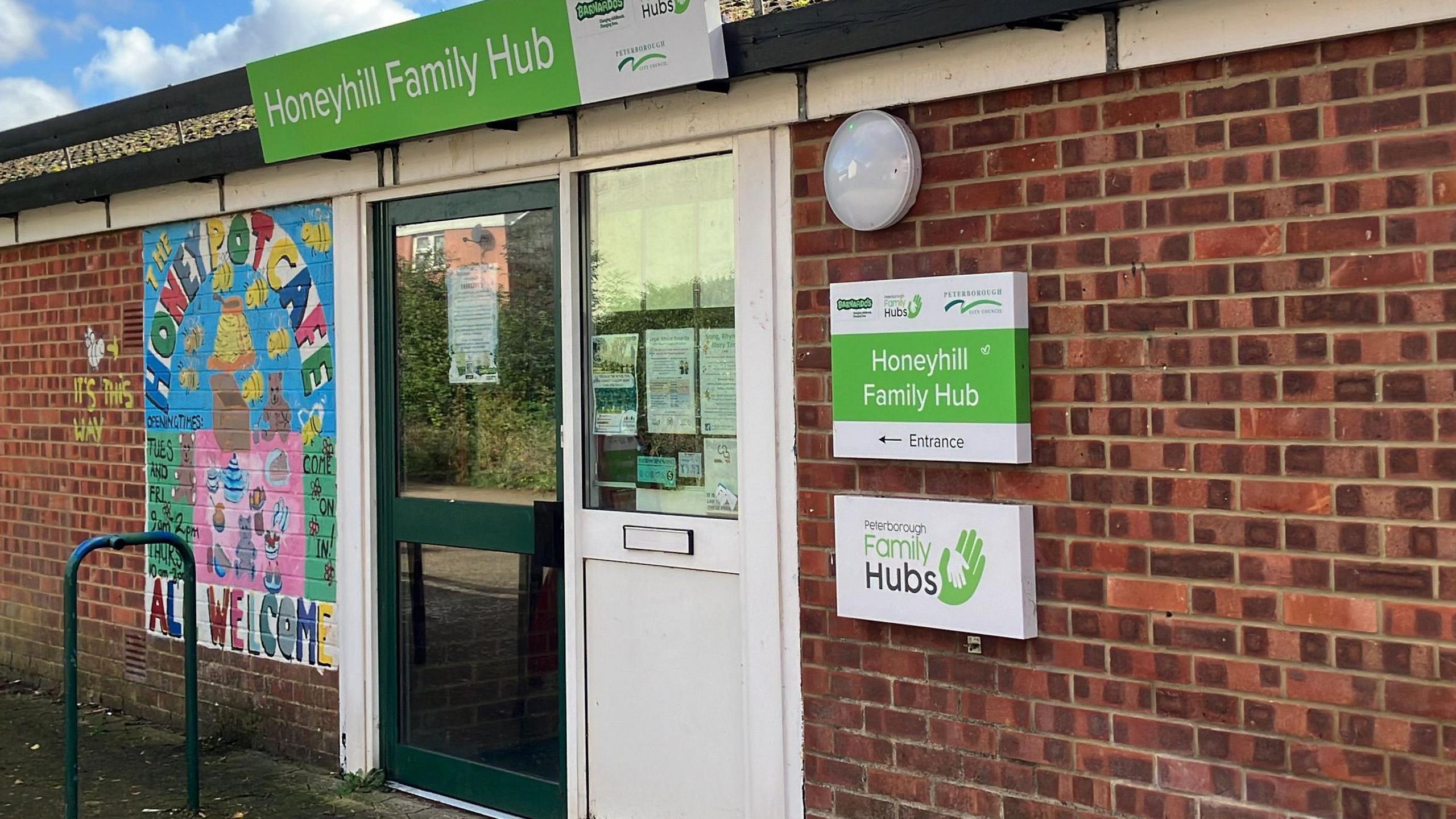 Honeyhill Family Hub, Peterborough. It is a single storey brick-built, flat-roofed building. A green door in the wall has a sign saying in white on a bright green rectangle Honeyhill Family Hub.