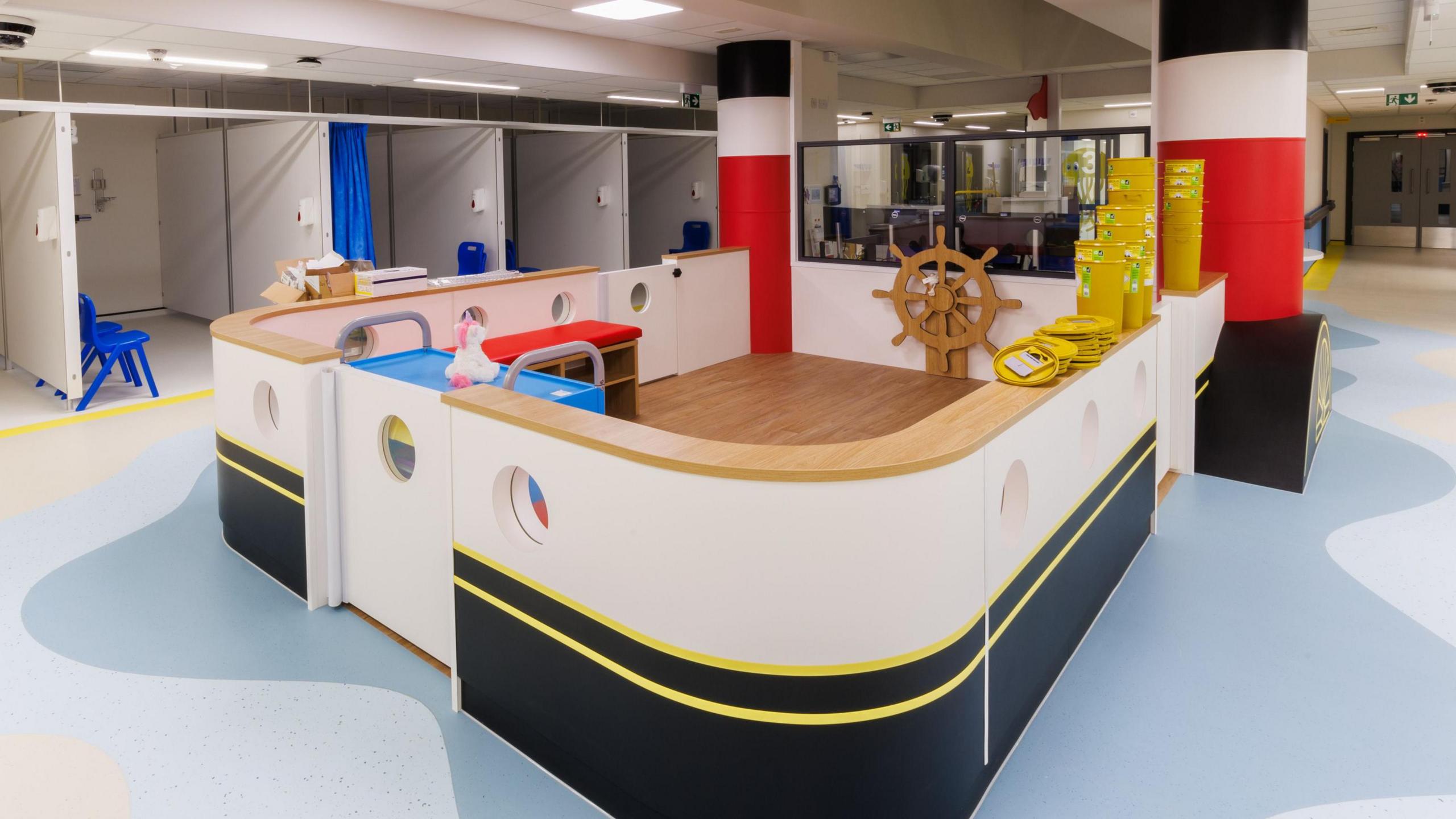 The children's emergency department at Queen Alexandra Hospital. It has a sectioned-off area painted like a ship with porthole windows and an old-wooden fashioned ship's wheel. To one side of the room are several small medical bays with blue chairs.