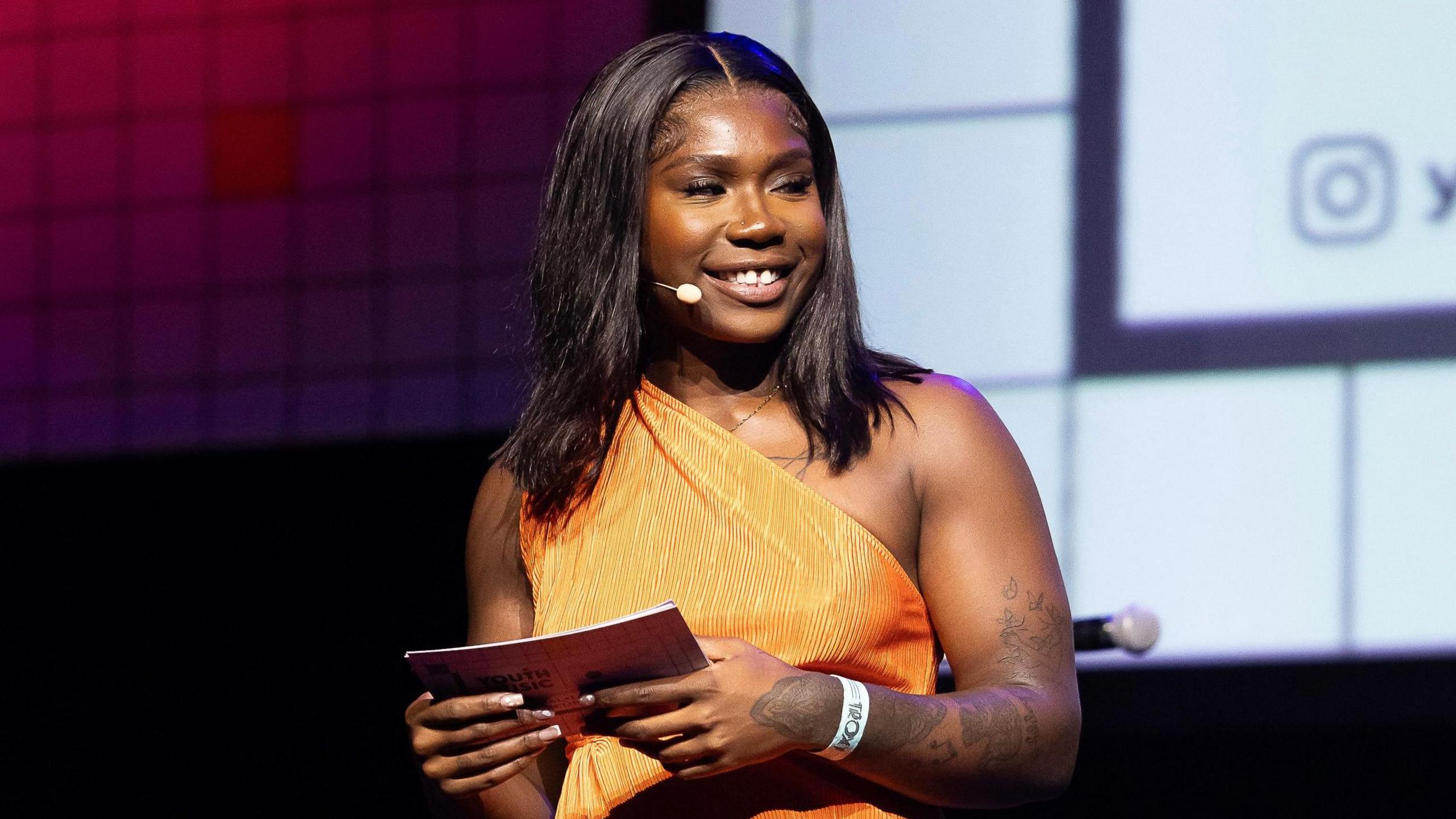 Jade Barnett in an orange dress standing on stage. She holds notecards and wears a microphone. She is at the Youth Music Awards 2024.