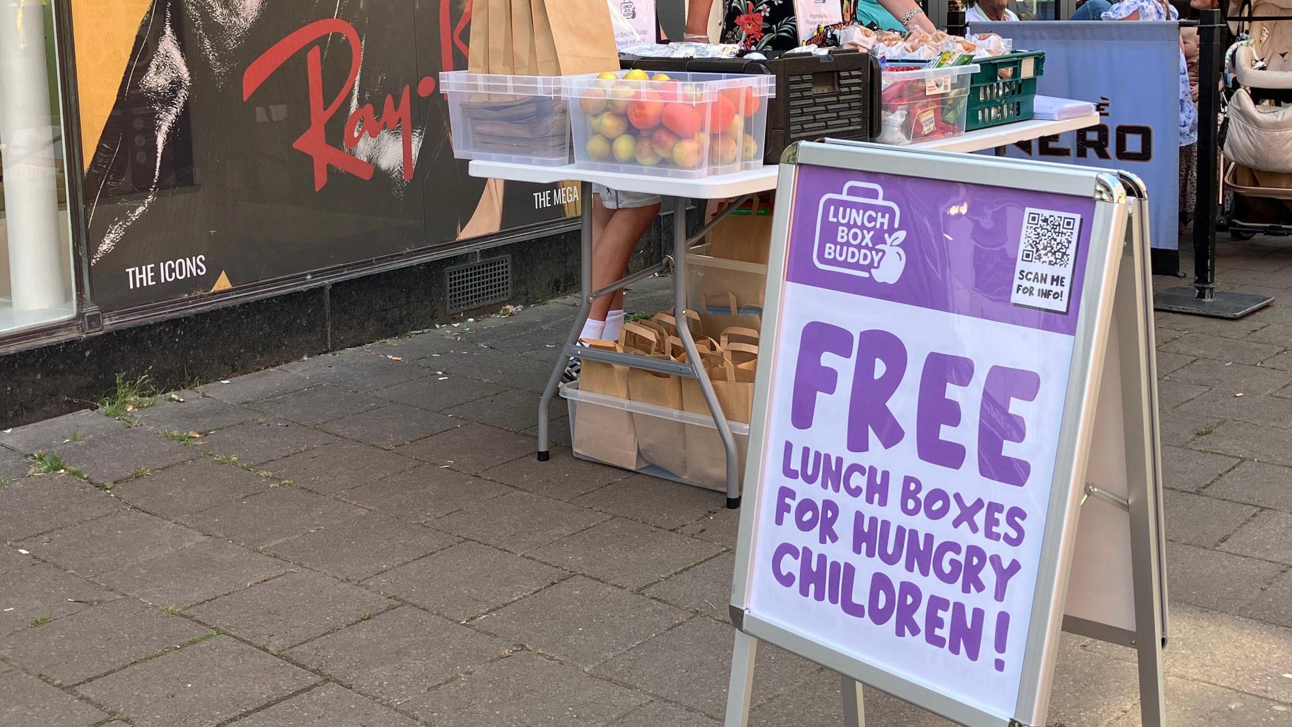 A pavement with a sign board which reads "free lunch boxes for hungry children" and has a QR code on top. Behind it is a table with see-through plastic boxes containing fruit and filled paper bags. A hoarding advertising sunglasses can be seen behind.