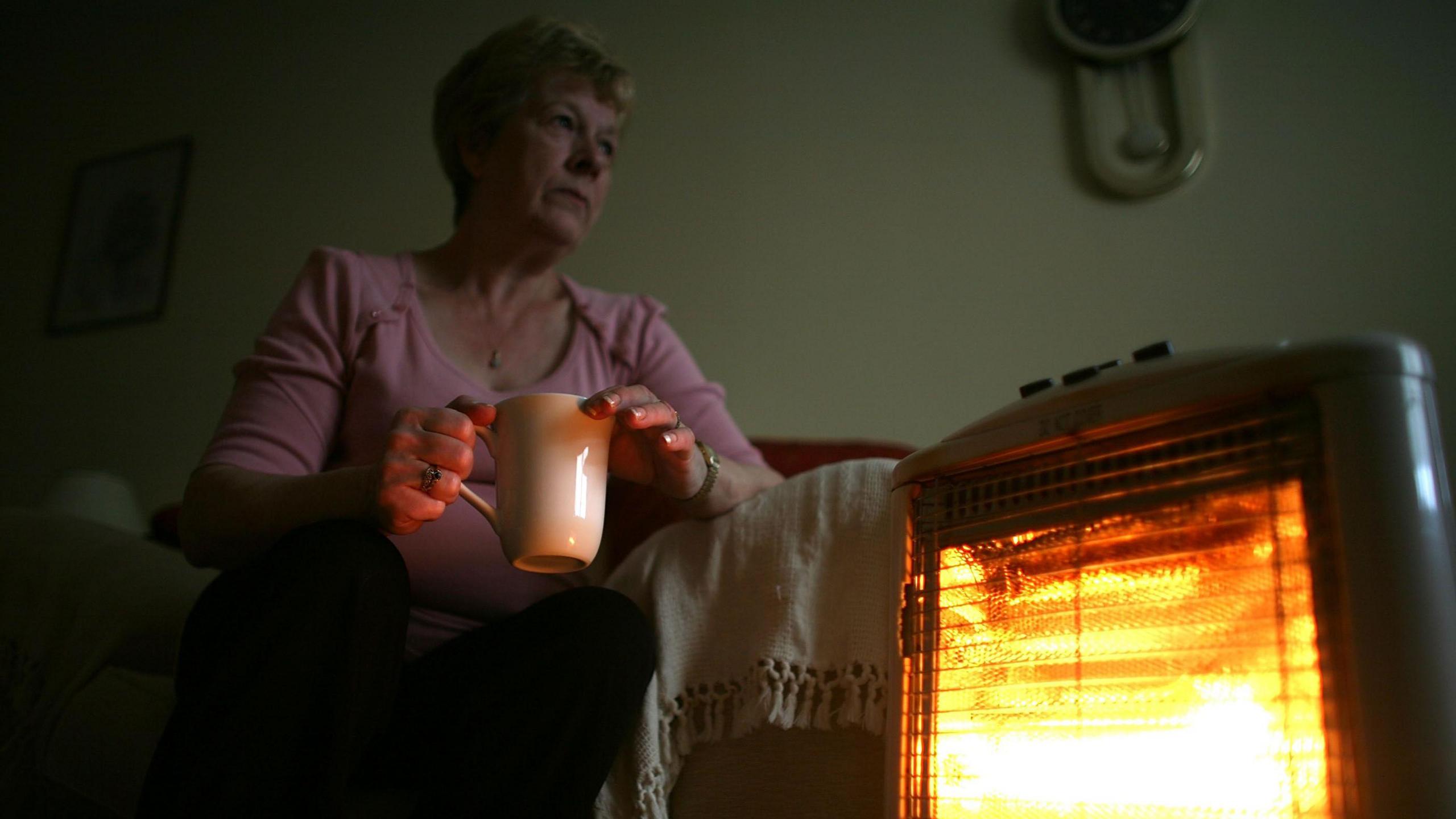 A woman sits by an electric heater in a living room. She is wearing a pink jumper and holding a mug.