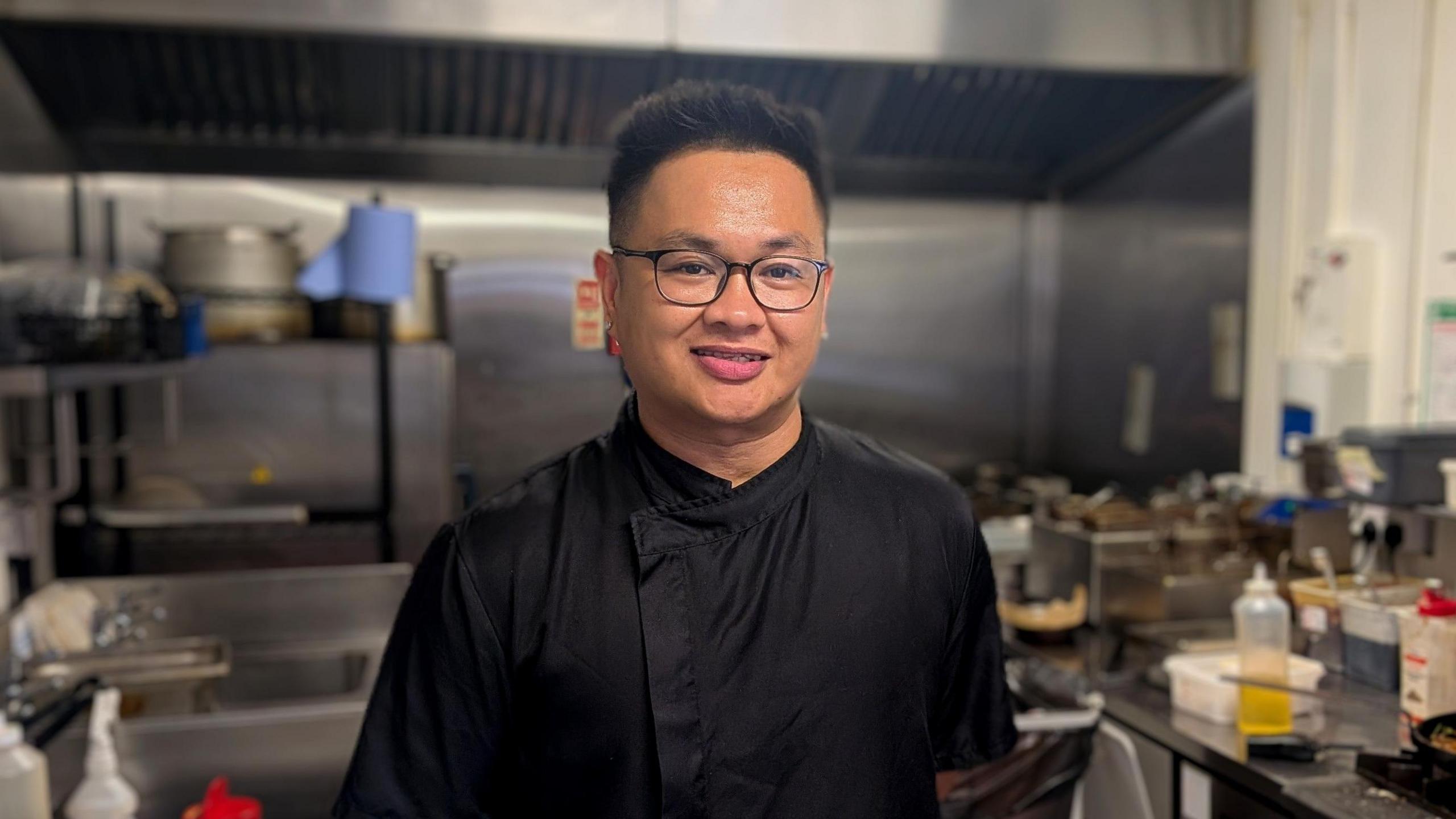Ryan Albano smiles into camera in a black chef's uniform. He has dark hair and black glasses and is standing in a commercial kitchen surrounded by cooking utensils and ingredients.