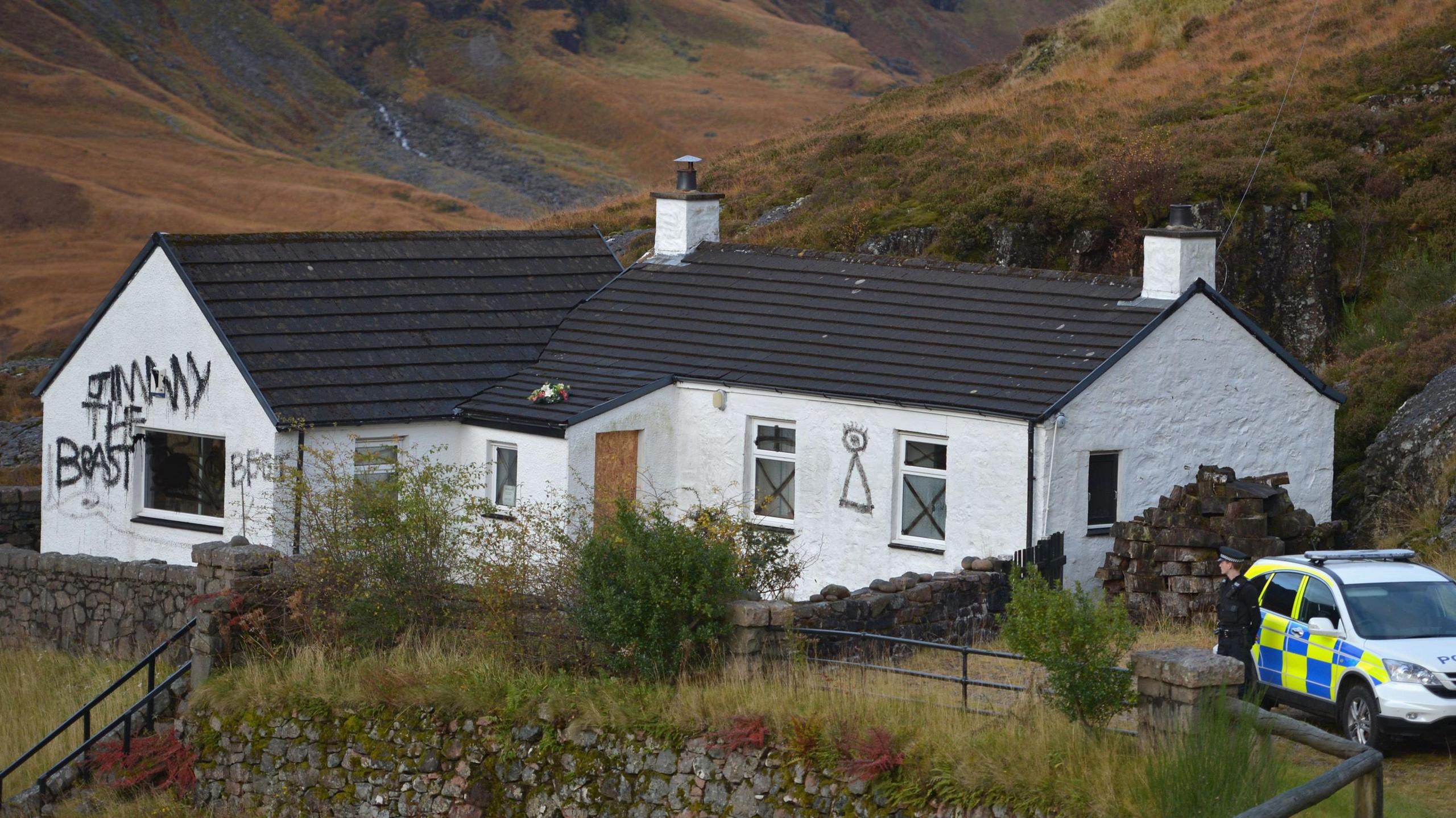 Exterior of the main cottage at Allt Na Reigh taken in 2012 - a white single story building with a black roof with spray painted graffiti on the walls. A police vehicle and officer stands outside.  