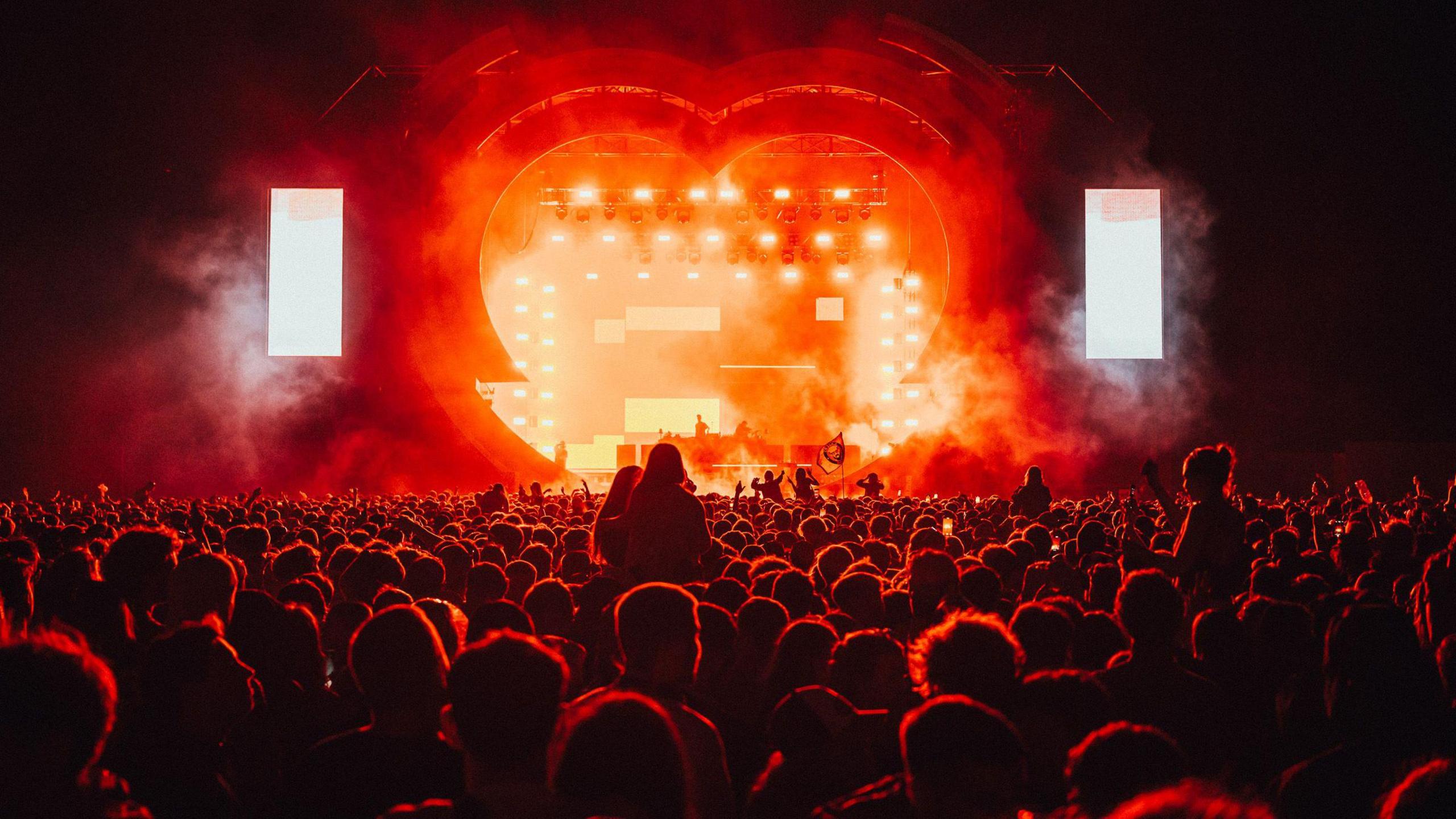 A large crowd is seen facing the stage at Love Saves The Day in Bristol. It is night time and the stage is in the shape of a heart and is illuminated by orange lights