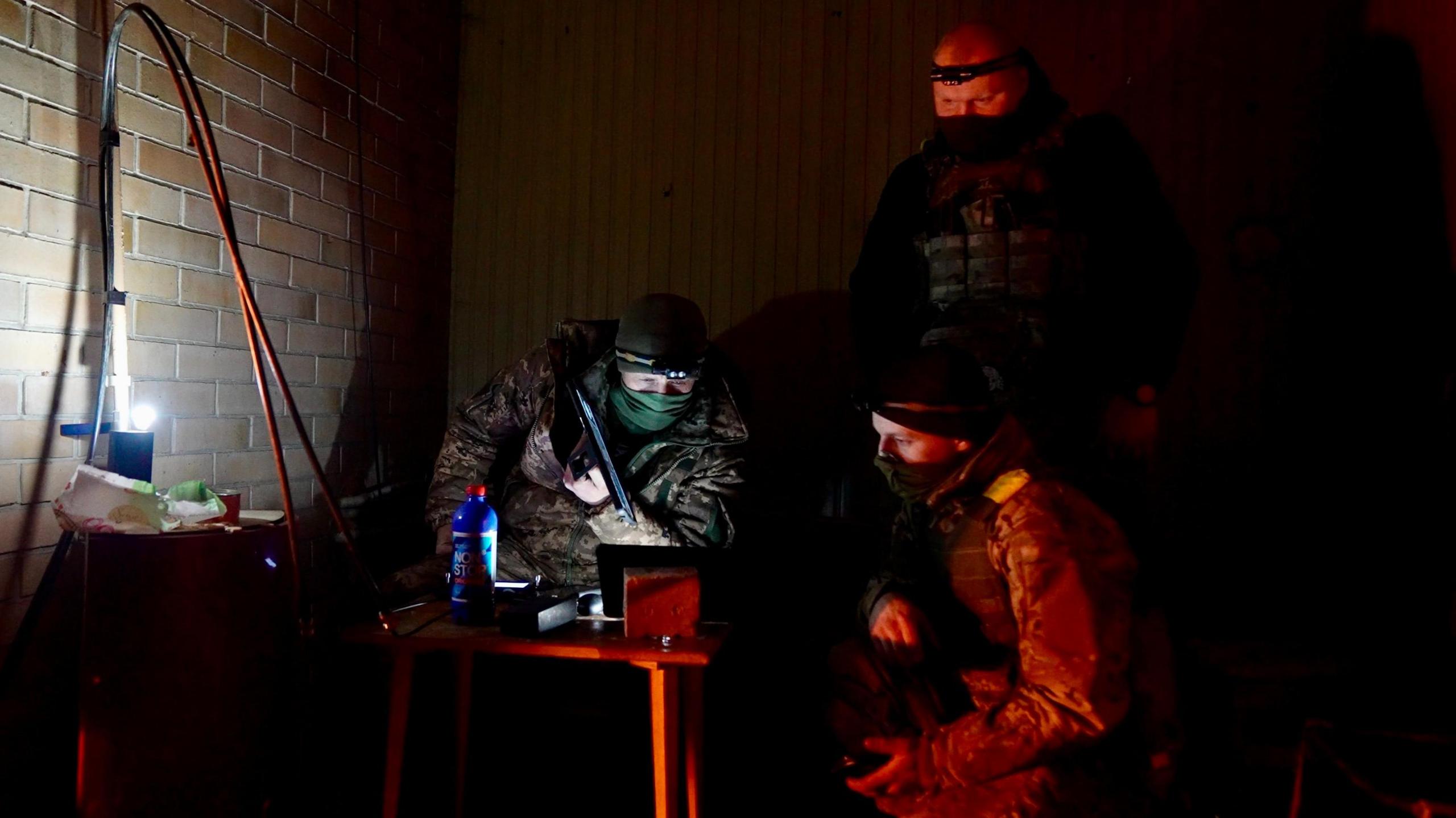 Two masked men look at a computer screen with another masked man standing over him, in a dark room lit up by one lamp