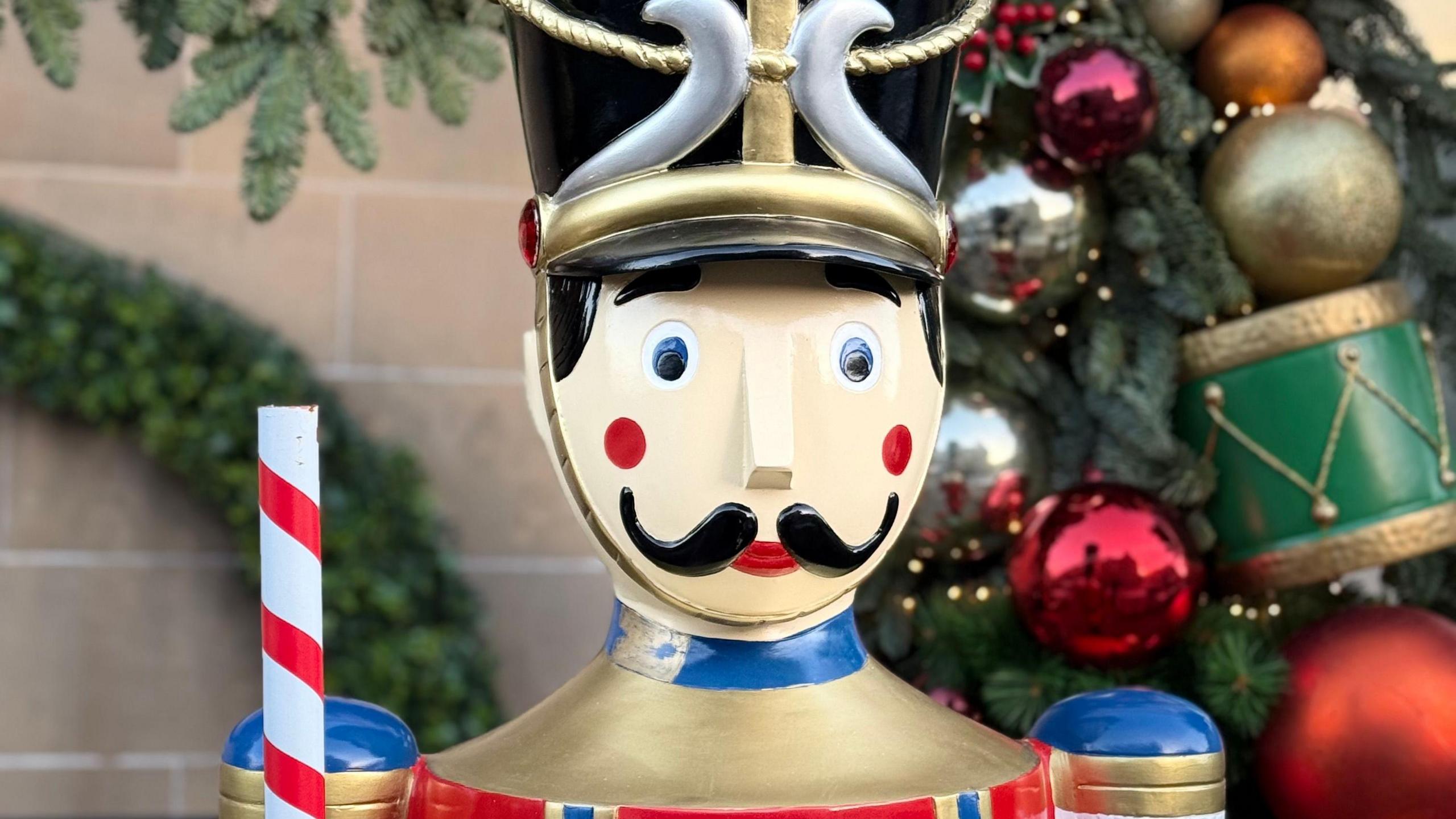 The statue has a black hat. It has a black moustache and red dots to give it rosey cheeks. It is holding a red and white stick. This is a head and shoulders shot so you can't see the body.