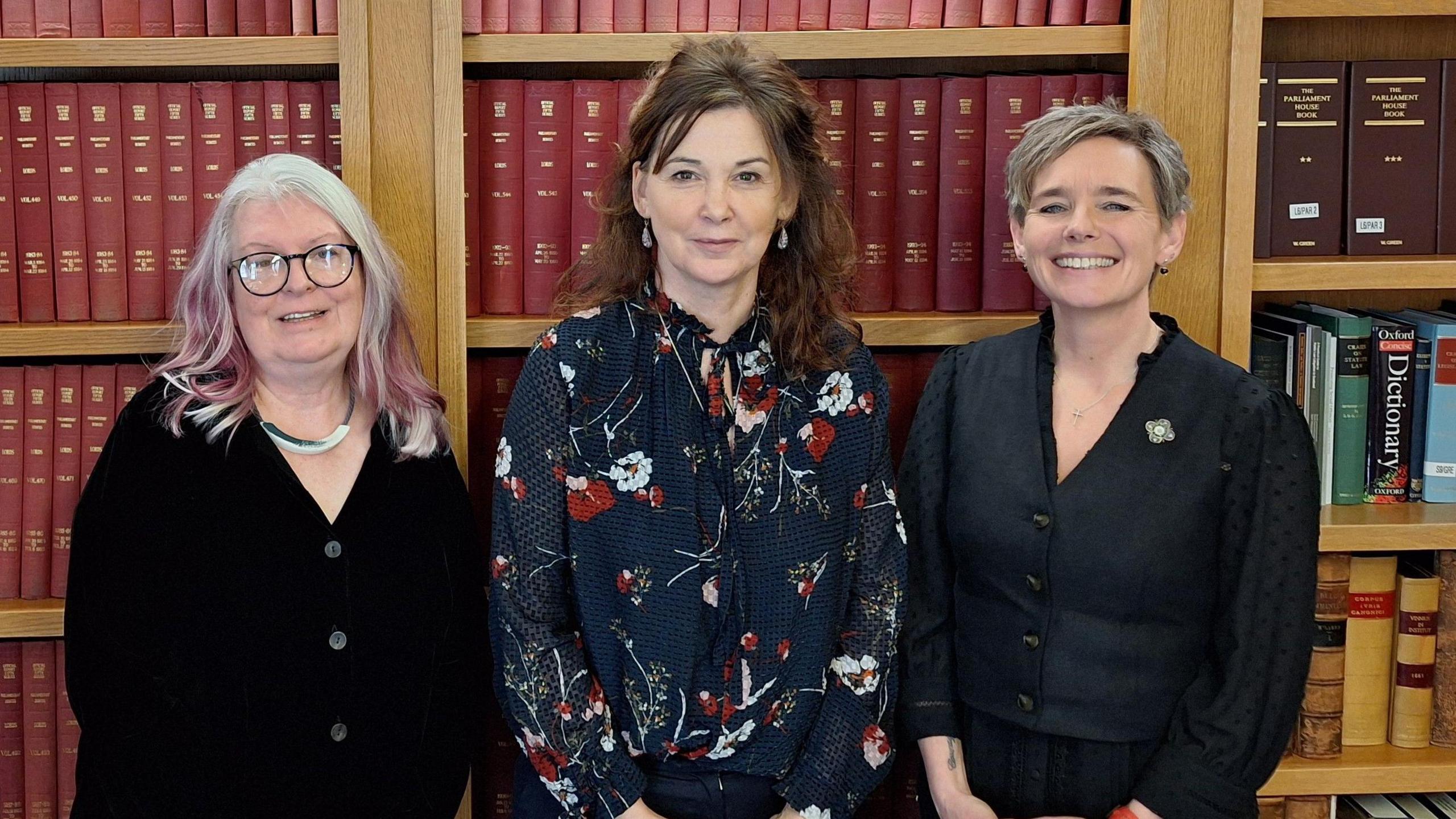 Suzy Angus with the Lord Advocate Dorothy Bain KC and Emma Bryson. They are standing in front of book shelves. Suzy has long white and pink hair and is wearing a black buttoned cardigan. Dorothy Bain wears a navy blue blouse with red and white floral print. Emma has a dark blue blouse.
