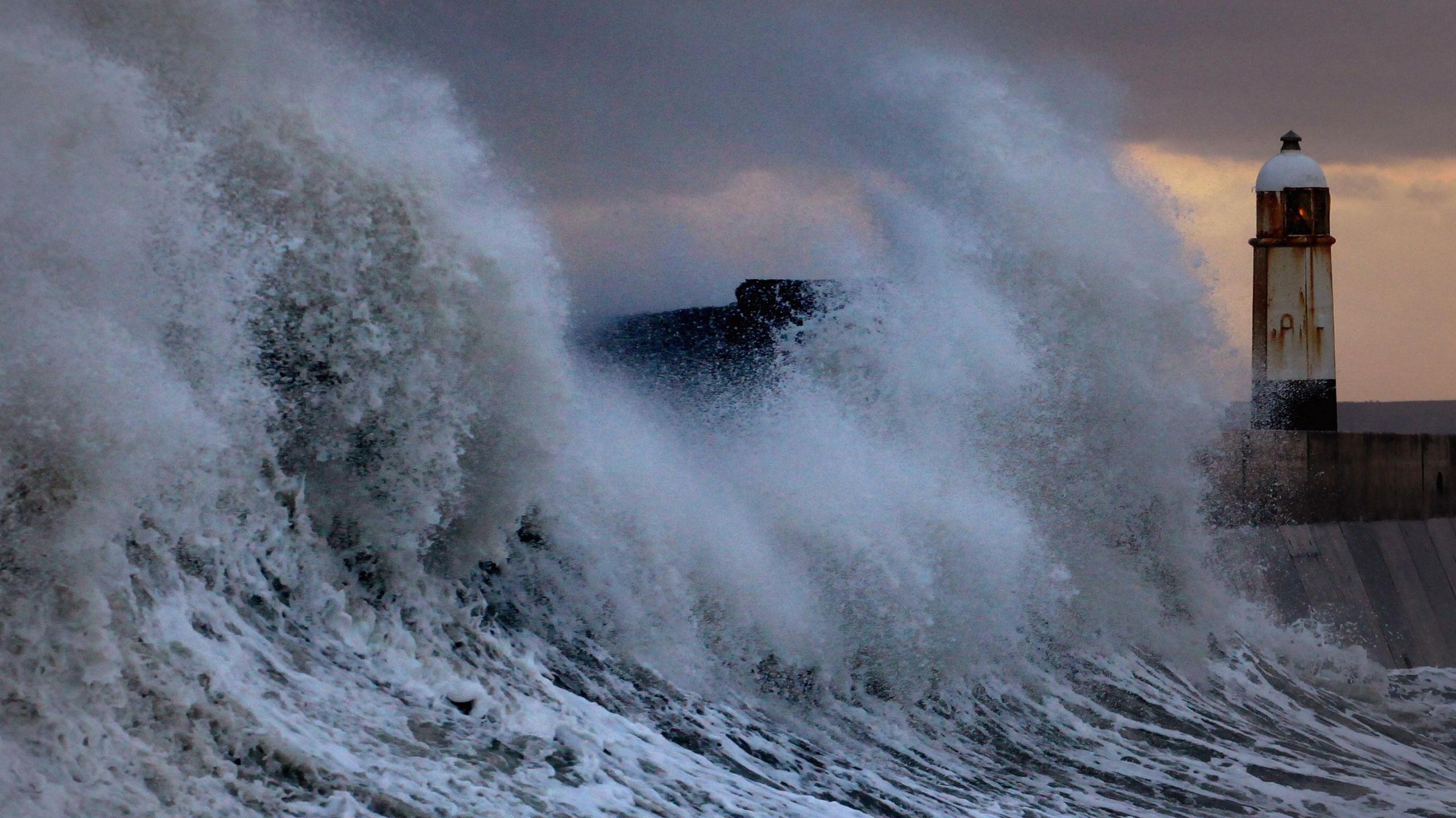 Waves crash over a harbour wall next to a lighthouse