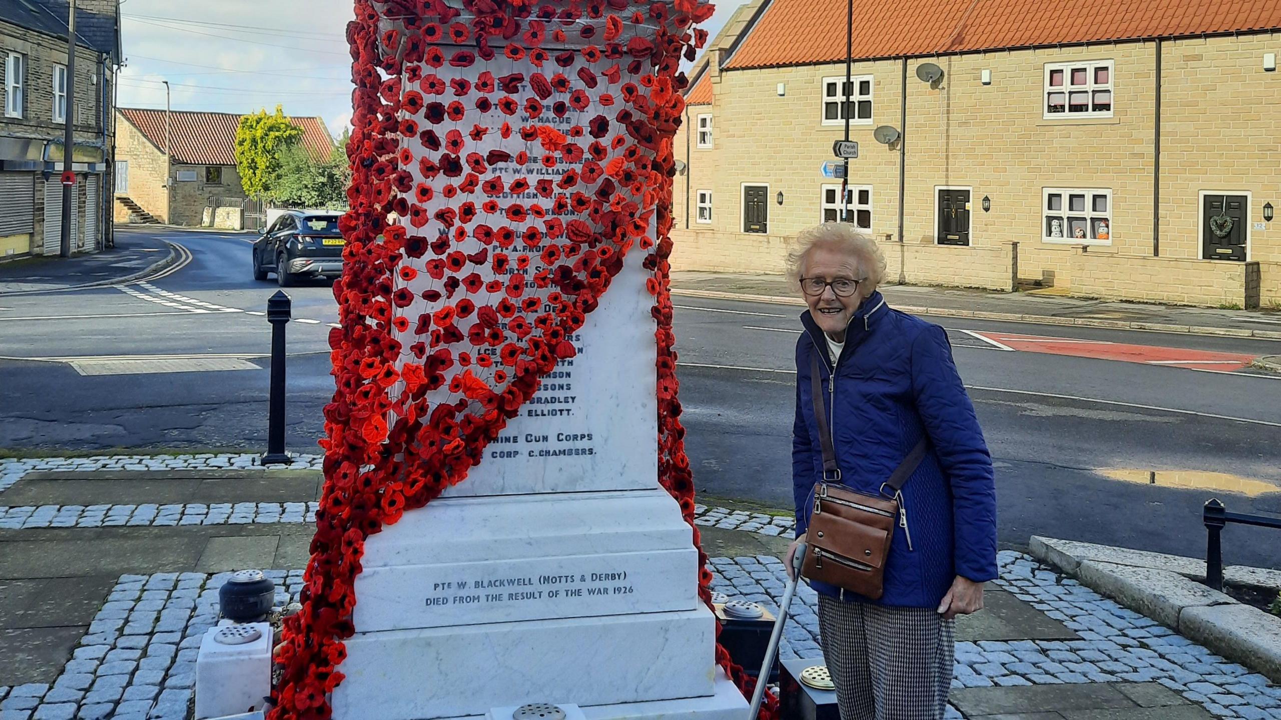 Betty Quinton is stood next to the cenotaph, in the parish of Wales. It's made of marble, with a bust of a British soldier on top of a tall podium. Names of local men who died during the First World War are engraved on it. At the bottom is a new inscription, dedicated to Walter.