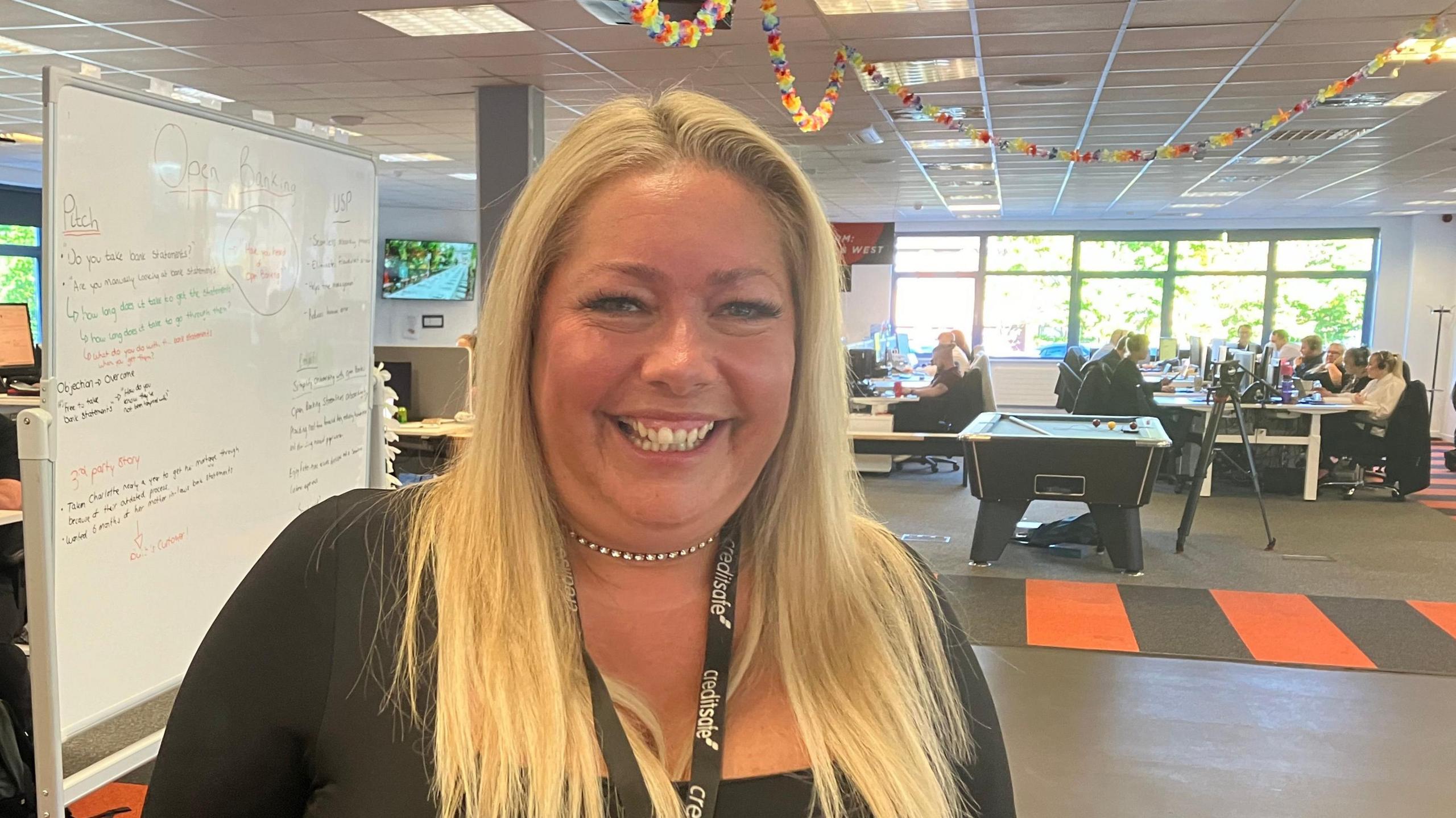 Natalie Brain, smiling at the camera, in the office, with workers at desks in the background, along with a whiteboard