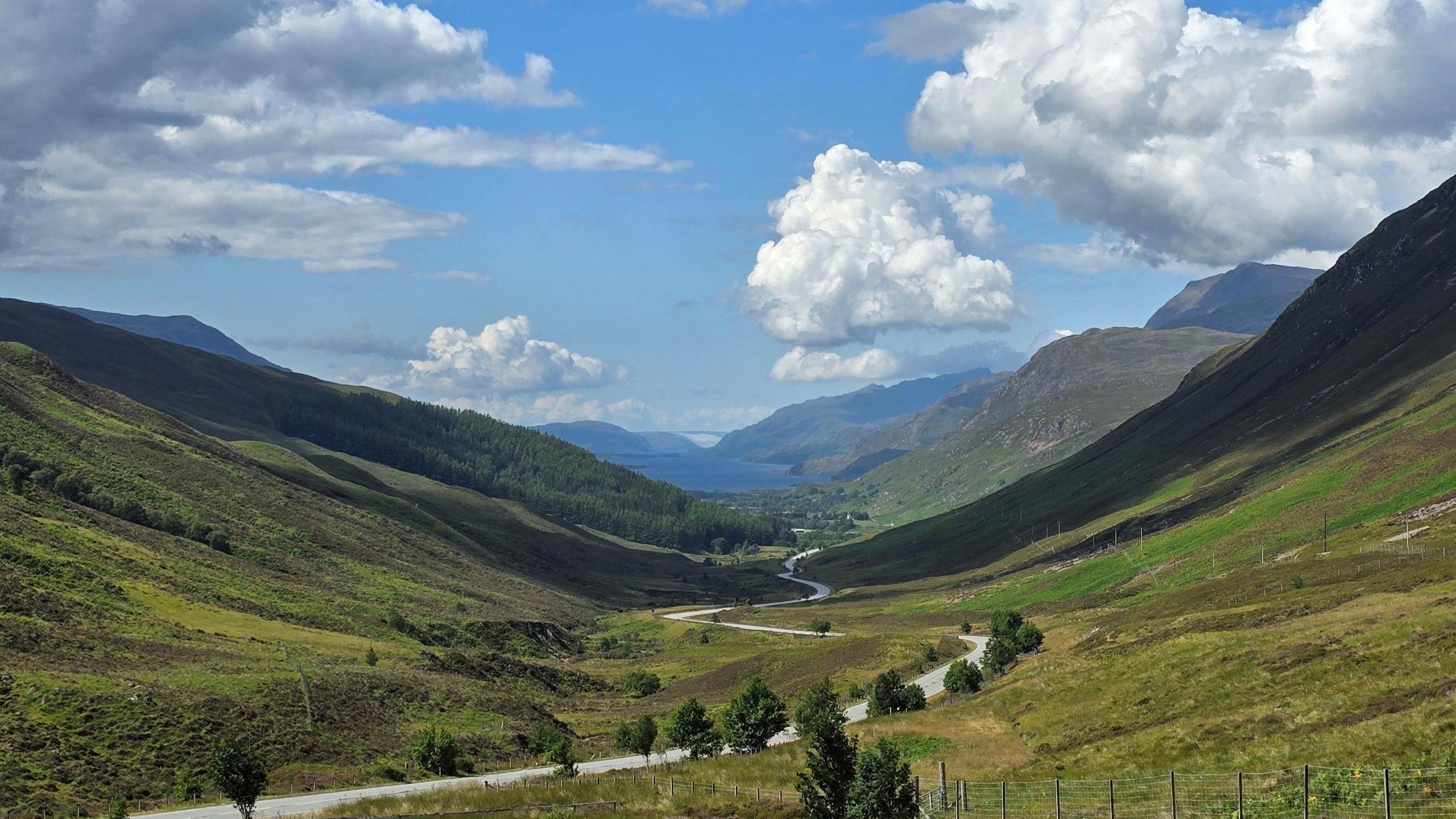 A valley surrounded by green mountains under a cloudy blue sky. A winding road runs through the valley with trees on either side. There are more mountains in the distance which are partially shadowed.