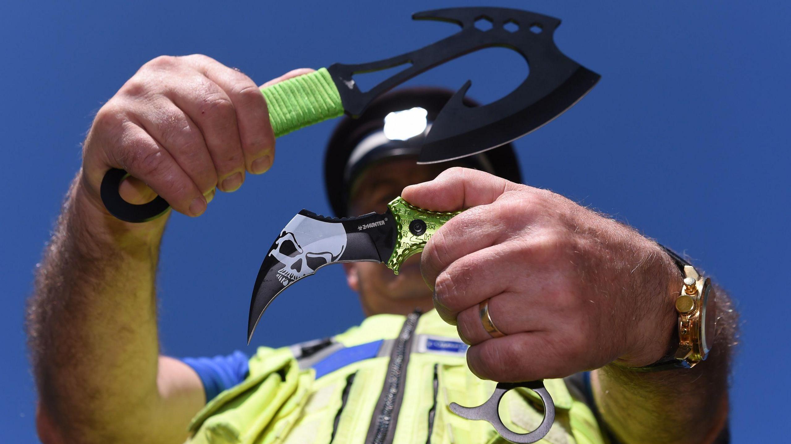 A police officer holds two 'zombie knives', which are curved blades with green handles.