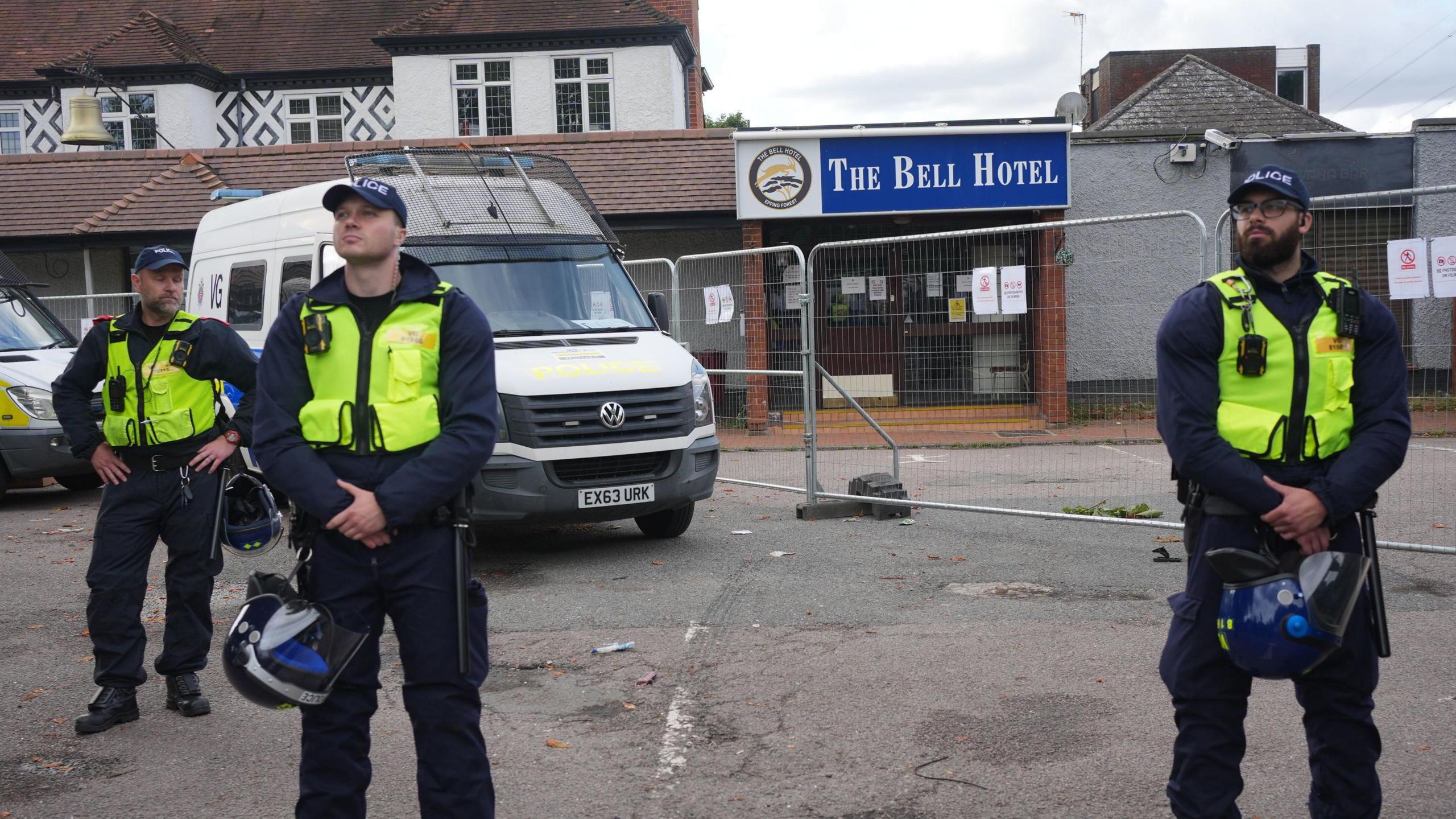 Three police officers wearing hi-vis vests standing in front of vans and the hotel, which has metal fencing in front of it.