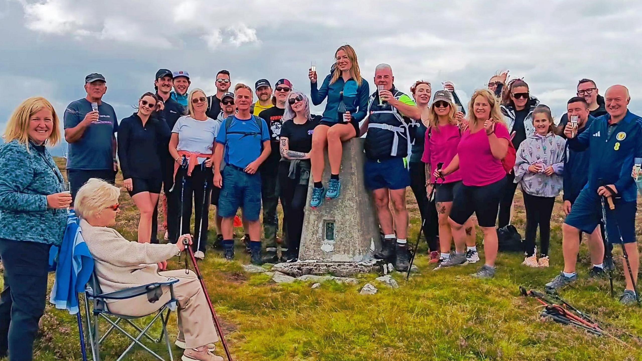 A group of around 20 people pose around a concrete trig point on a grassy hilltop under cloudy skies. Several people hold walking poles and drinks, while one person sits in a folding chair in the foreground. The landscape stretches into the distance behind the group, suggesting a hillwalking or summit gathering.