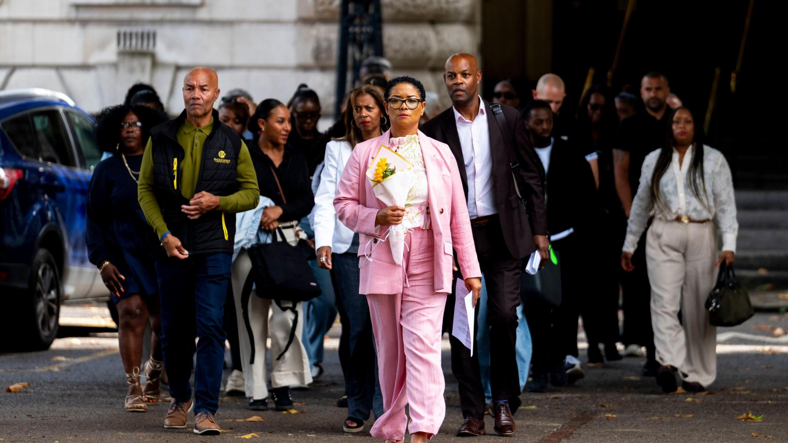 A group of people outside a court building, a woman at the front in a pink suit, holding a bunch of flowers.