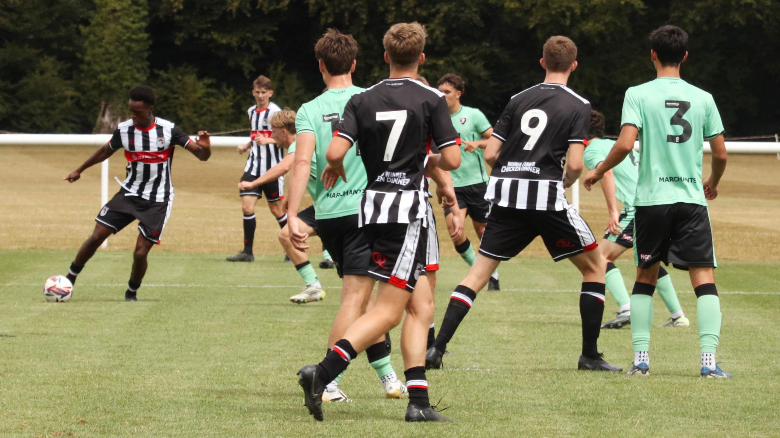 A group of men playing football on a pitch. None of them are looking at the camera. They are all in kit - four of them are wearing green t-shirts and four are wearing black and white striped t-shirts. 