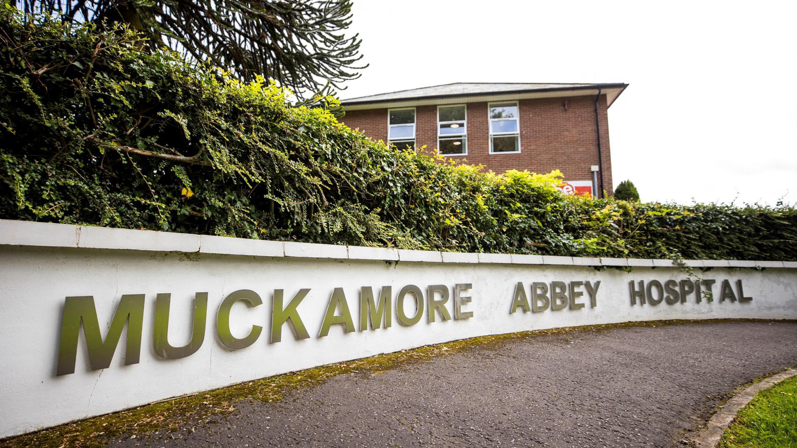 A white wall reads 'Muckamore Abbey'. There's a red brick building behind the low wall.  A small green bush runs along the top of the white wall. 