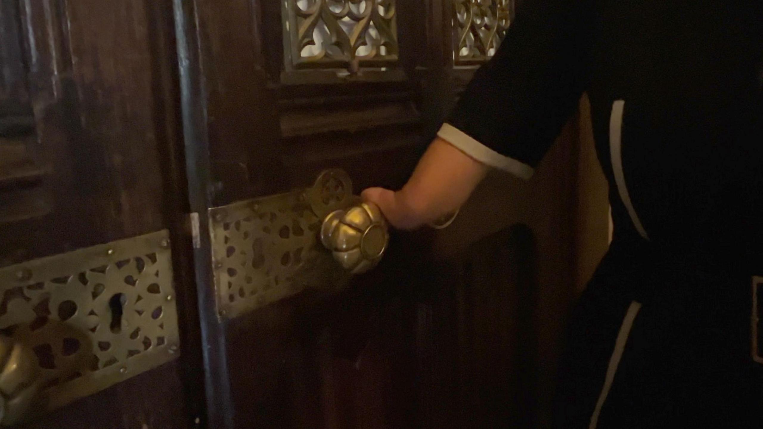 A woman's hand, with only one digit, on a gold-coloured round doorknob. The doorknob is smooth and shaped like a flower with petals. The door is dark wood with metal decoration over the window and around the doorknob plate. The woman is wearing a dark dress.