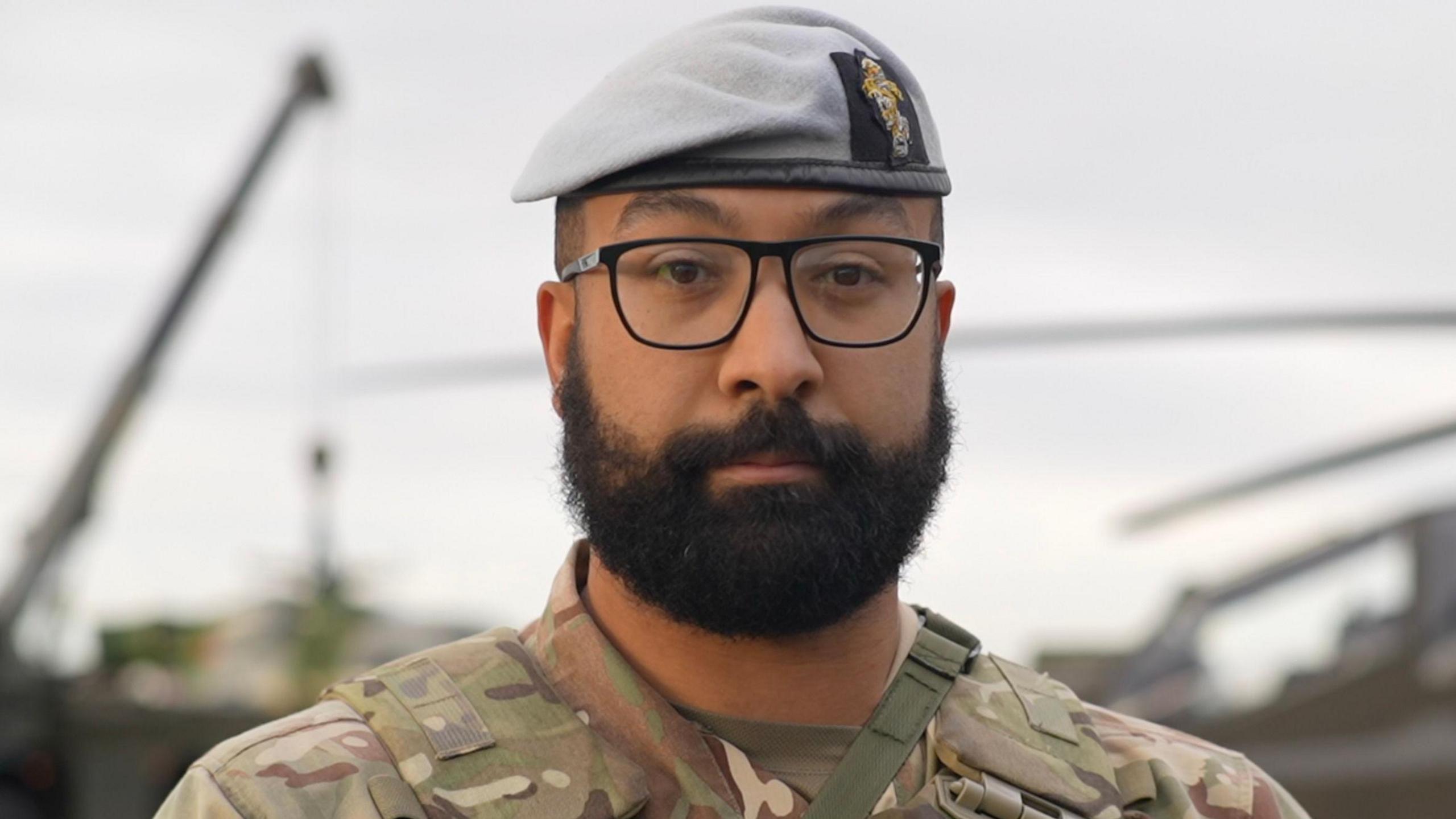 Blake smiles at the camera. He wears a grey beret hat, glasses, camouflage uniform and he has a dark thick beard.
