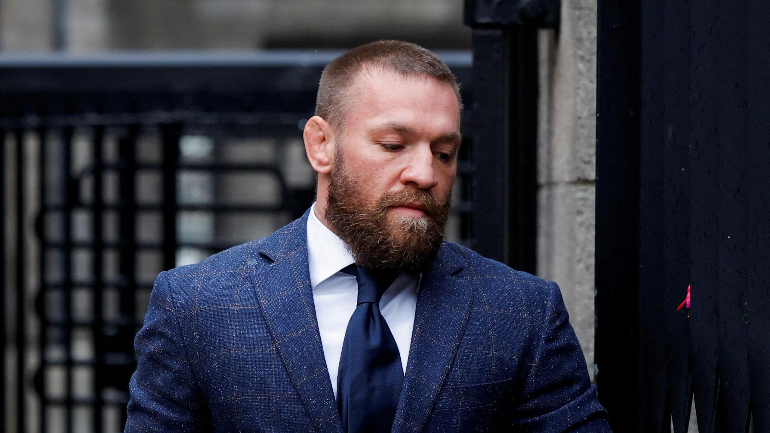 Conor McGregor has short hair and a beard. He is wearing a blue jacket, blue tie and a white shirt. Behind him is a security gate at the entrance of a court building.