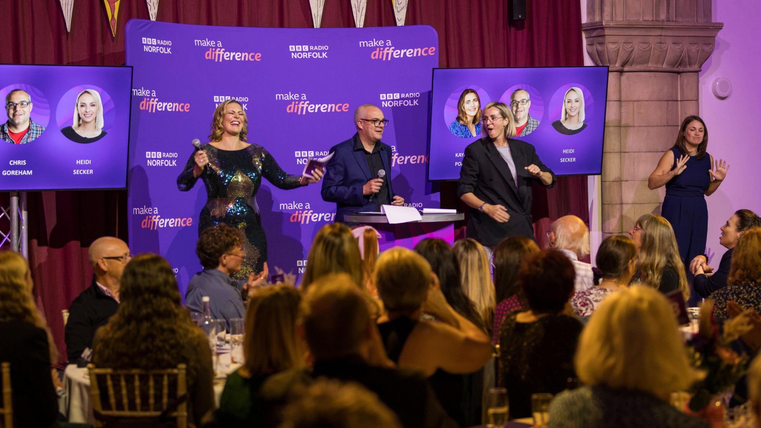Two women and a man stand on stage in front of a purple back drop with two screens. They are in front of an audience.