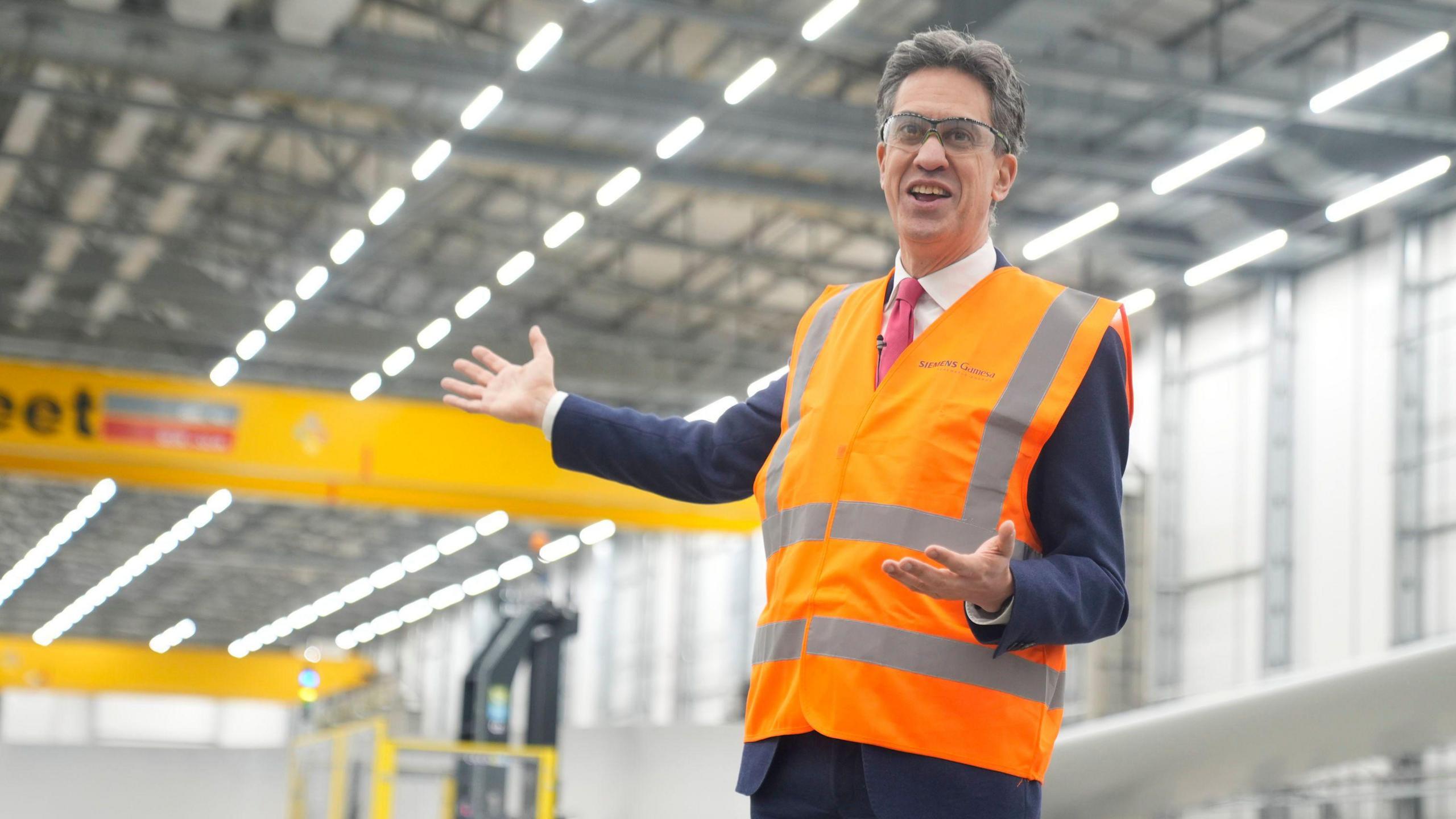 Energy secretary Ed Miliband stands smiling inside a large, modern-looking factory. He is wearing an orange and grey hi-vis jacket.