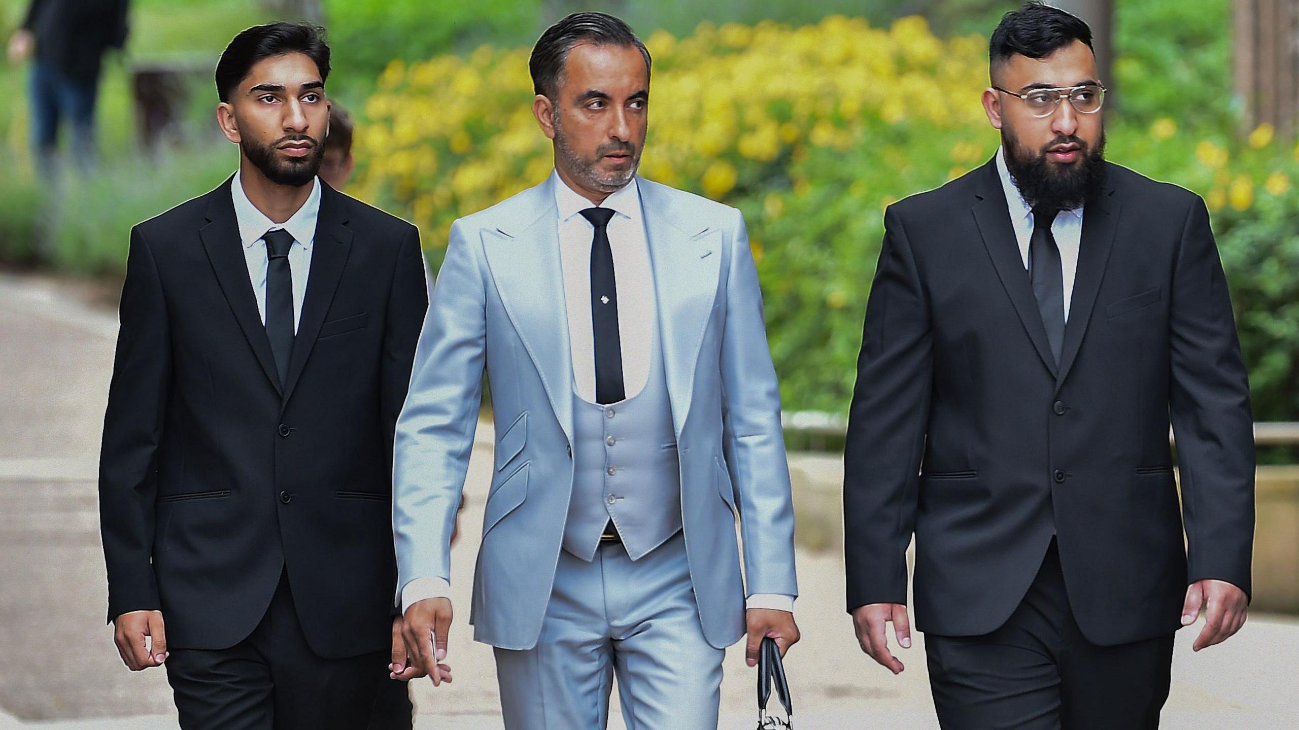 Two young men wearing matching black suits and ties walk along a street on either side of an older man. He is wearing a light blue three-piece suit and carrying a briefcase. They all have serious expressions.