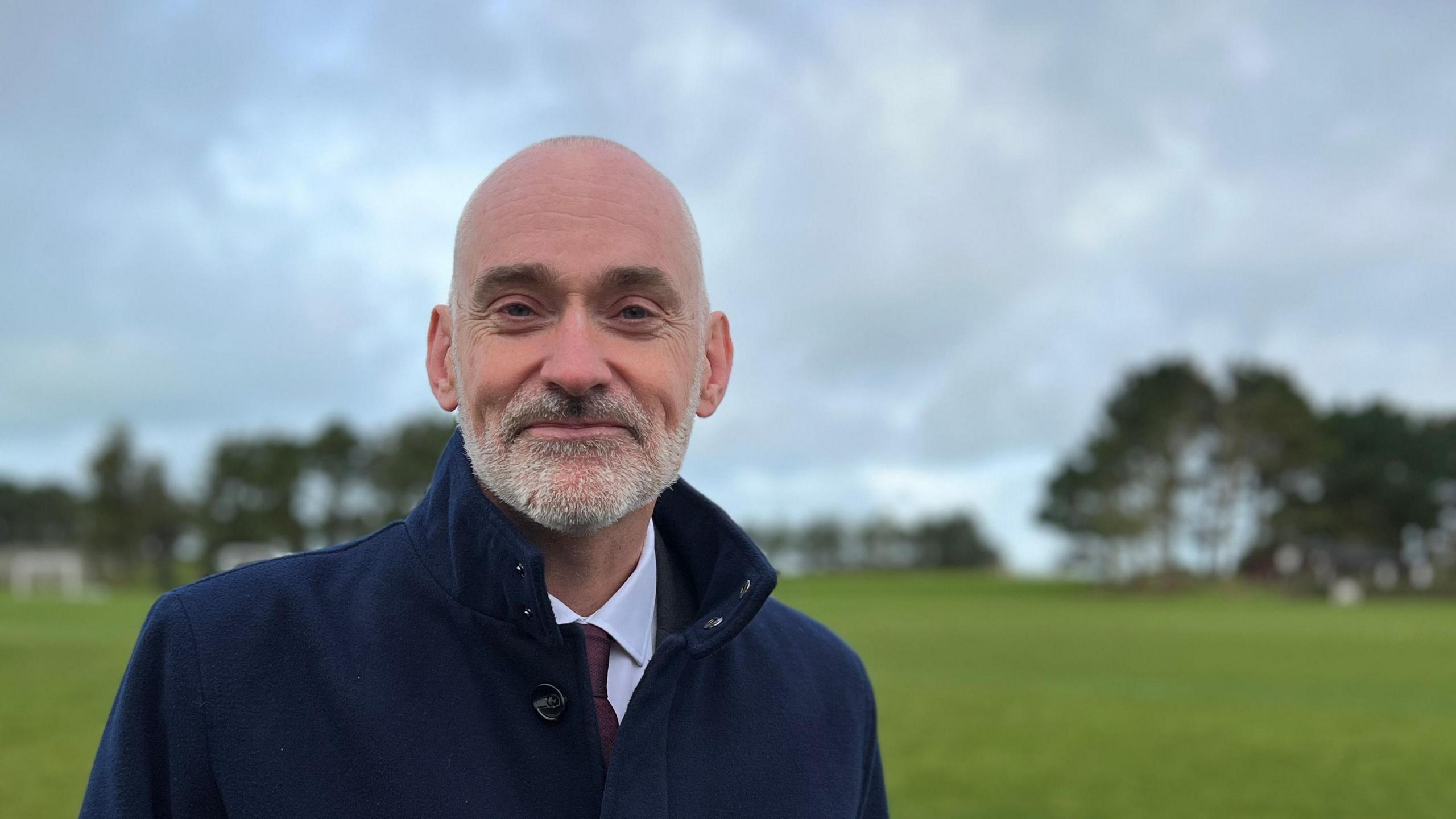 Peter Bradley. He is wearing a blue jacket over a white shirt and maroon tie. He is bald with a short grey beard. He is stood on some grass with trees in the background. 