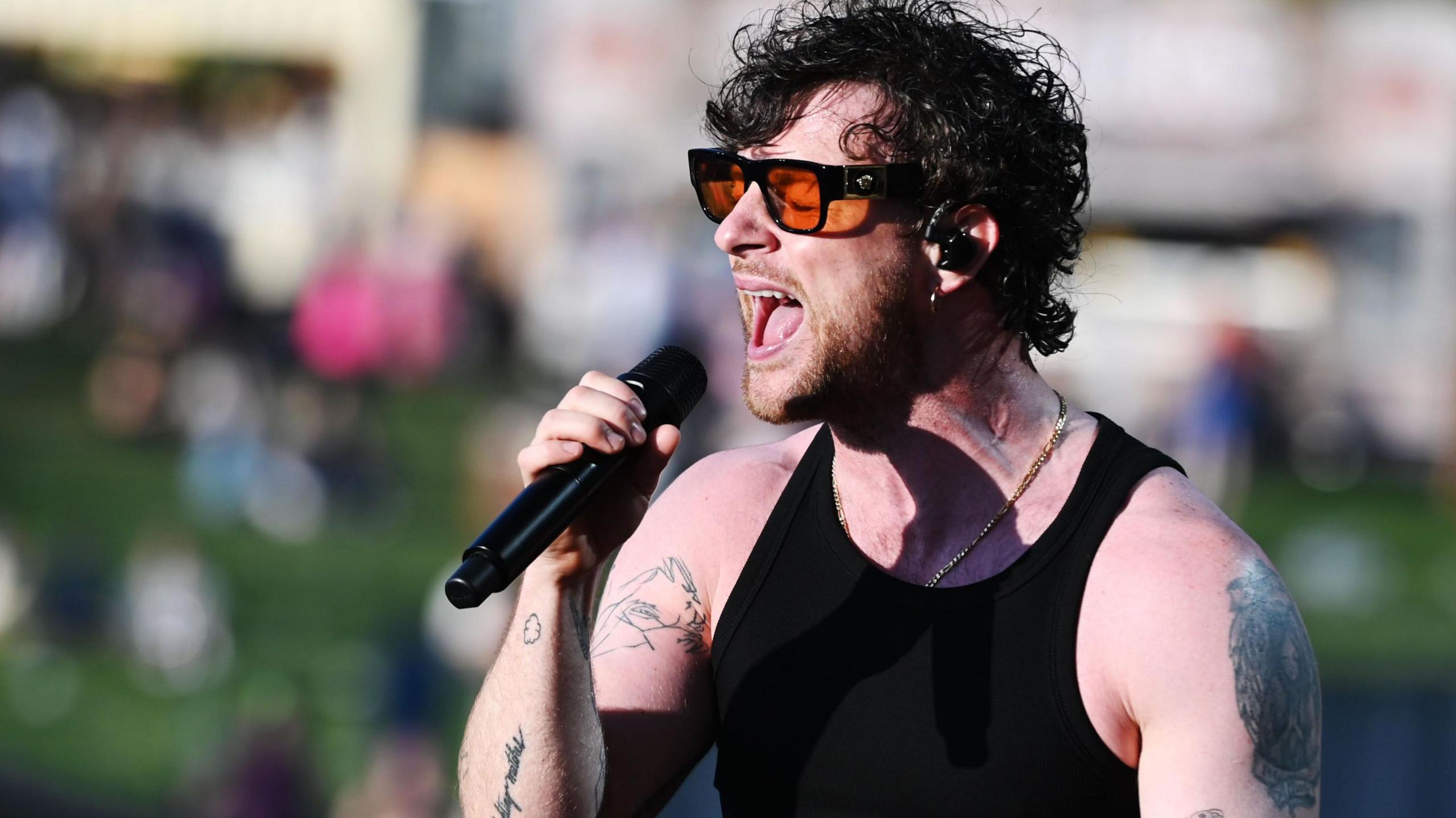 Tom Grennan, wearing orange-tinted black glasses and a black vest top, sings into a microphone at Radio 1's Big Weekend in 2023