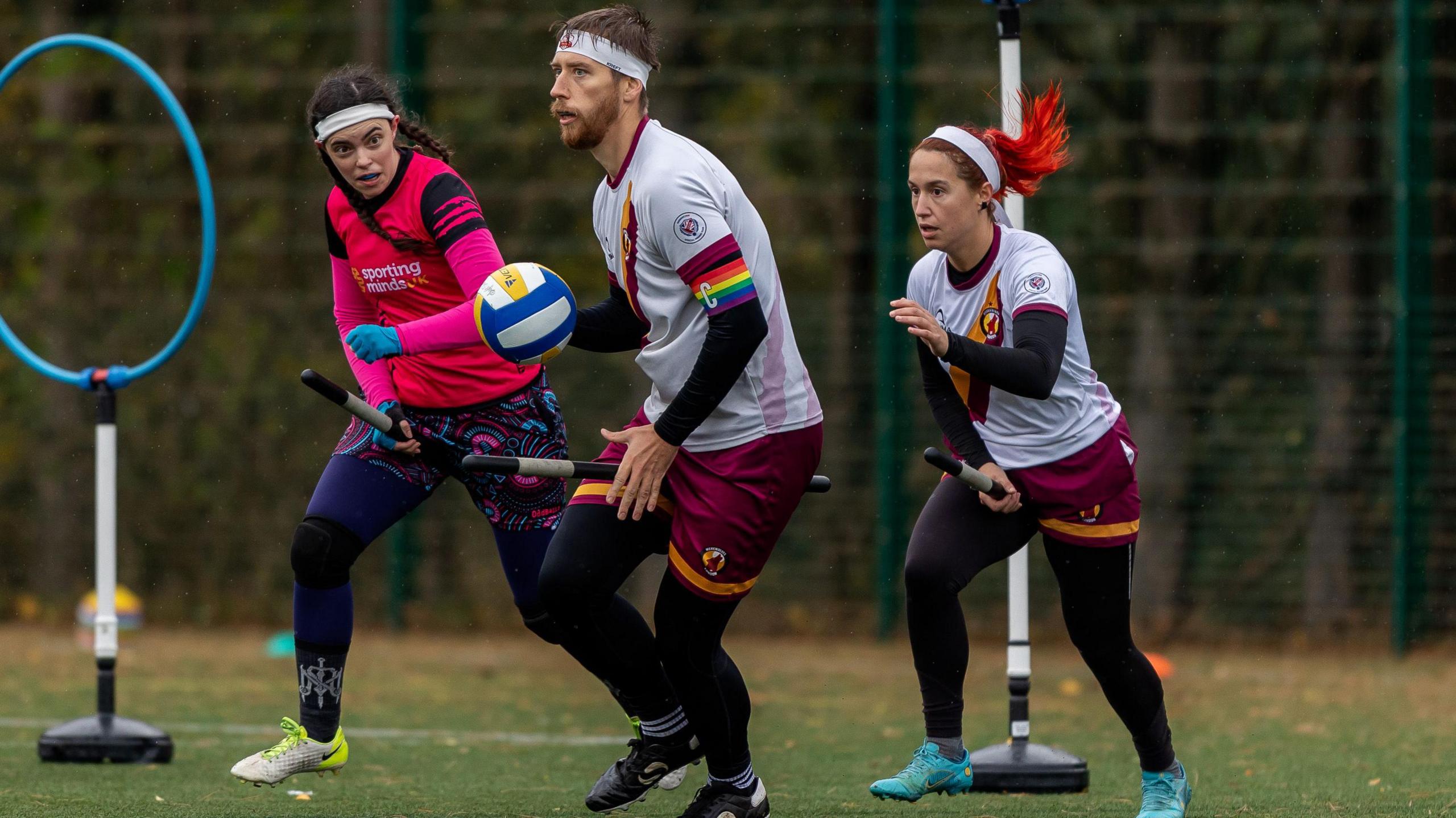 Three people dressed in sports kit play in front of two hoops fixed to metal poles. One player holds a ball, while two others run next to them. All players hold short metal poles between their legs.