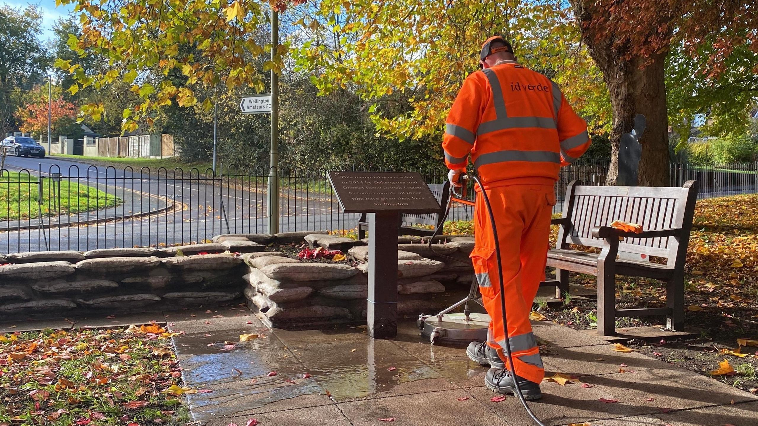 Mike pictured using a large, industrial pressure washer on flagstones next to the memorial, which is a cross-shaped flower bed with a bench next to it and a plaque. He's wearing a bright orange hi-vis jumper with silver reflective stripes, and a bright orange baseball cap. He's stood in a park, with the grass covered in orange leaves, and the leaves still on the trees are shades of red and orange. There's strong sunshine and the sky is blue. 